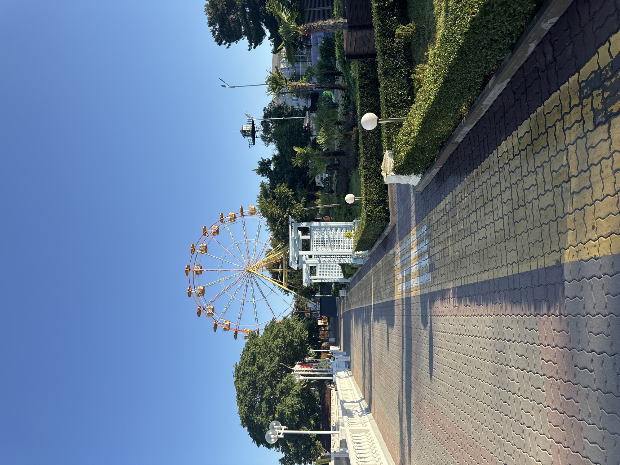 A pedestrian walkway with a patterned cobblestone surface leads towards a Ferris wheel in the background. The walkway is bordered by a white balustrade on the left and manicured green hedges on the right. Several lampposts and trees are visible along the walkway, as is a decorative white gazebo structure. The Ferris wheel is a prominent feature, with red and yellow cabins against a clear blue sky. Lush green trees and palm trees are interspersed throughout the scene, creating a park-like atmosphere. Sunlight casts shadows across the walkway, indicating it is daytime. There are no people visible in the image.