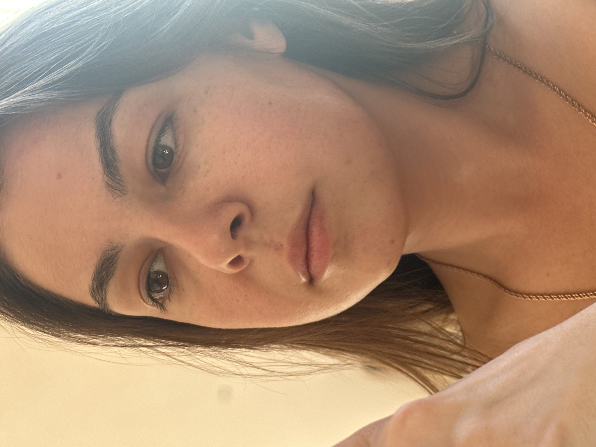 The image is a close-up portrait of a young woman's face, taken from a slightly low angle. She has long, dark brown hair that falls around her face. Her skin appears to have some freckles. She is wearing a thin, gold-colored chain necklace. The background is a plain, light-colored wall. There is a hint of a hand in the lower left corner of the image. The overall lighting suggests an indoor setting with natural light coming from the right. There is no discernible location in the city of Gyumri, Armenia.