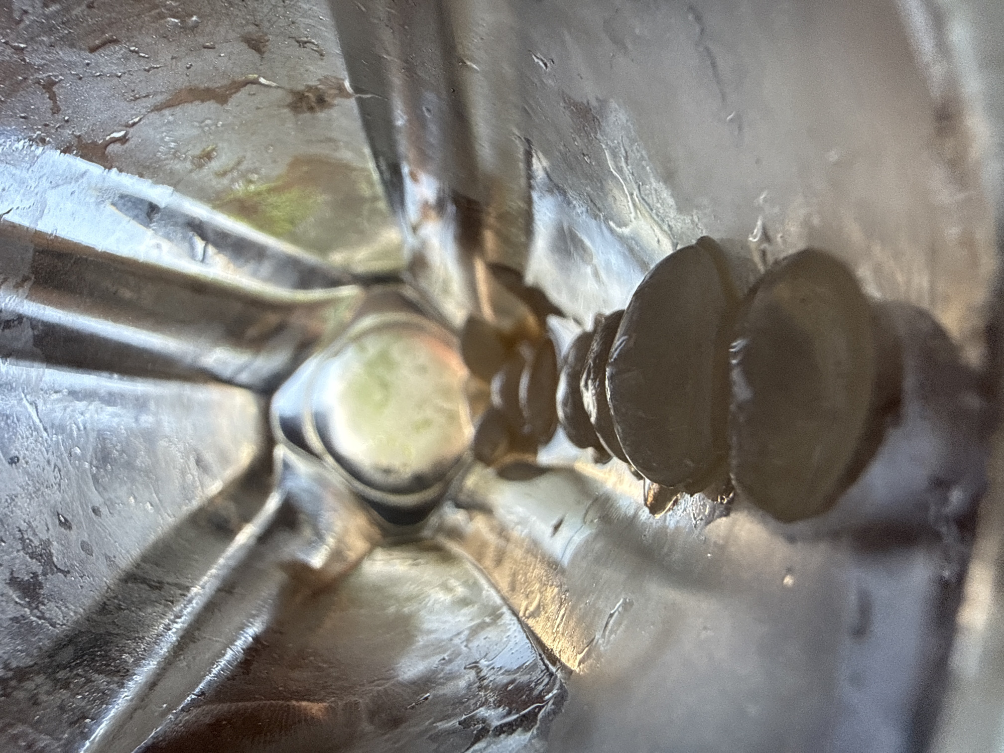 The image captures an interior perspective within a reflective, ridged, metallic conduit or container. The view extends longitudinally, from a wider foreground opening towards a distal, bright, circular aperture. Along the base of the structure, a linear arrangement of at least five dark, biconvex, ovoid objects, consistent with large seeds (e.g., pumpkin seeds), is visible, extending from the immediate foreground into the mid-ground. The metallic interior surfaces exhibit vertical corrugations, pronounced specular reflections, and evidence of moisture, including droplets, streaks, and residue accumulation. The distal aperture reveals a blurred background illuminated by natural light, showing indistinct shapes and colors, including hints of green foliage. No individuals are present, and no discernible actions or interactions are occurring. The scene is located in Unknown, Unknown.