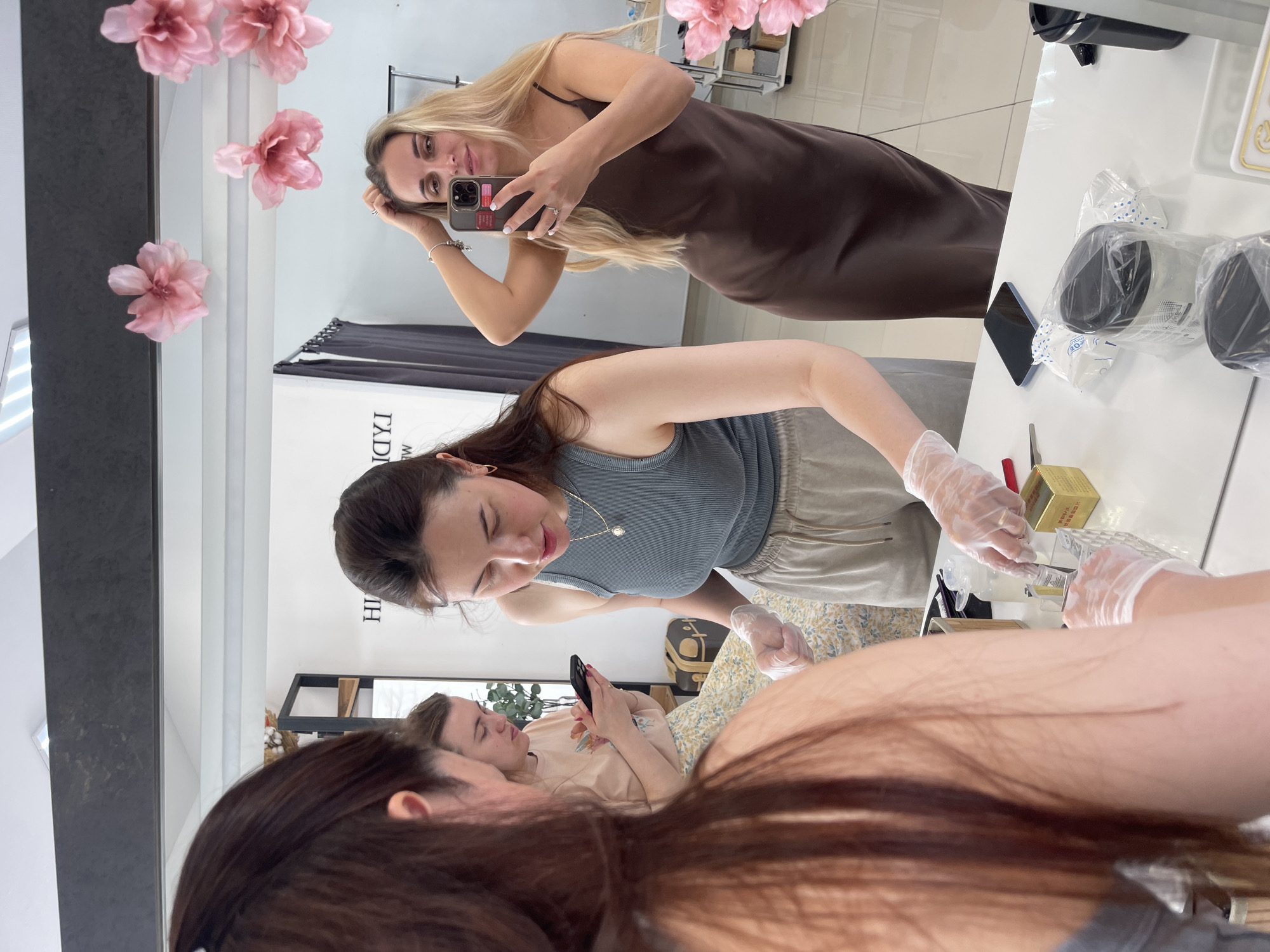 The image depicts a scene inside what appears to be a beauty salon or aesthetic clinic, possibly located in the city of Chaikovsky, Russia. A woman with long brown hair is visible in the foreground. She is receiving a cosmetic procedure, with her leg and arm exposed and being treated. Another woman, wearing a light gray tank top and light-colored pants, is administering the procedure. She is wearing gloves and appears to be applying something to the client's skin. A third woman is lying down in the background, looking at a phone. In the upper section of the image, a mirror reflects the scene. A fourth woman is holding a phone and taking a picture of herself and the scene, capturing herself in a brown dress. Various cosmetic supplies are present on a white table, including what appears to be a box of wax, applicators, and wrapped items. The environment seems clean and well-lit, indicating a professional setting. Pink artificial flowers are placed next to the mirror.