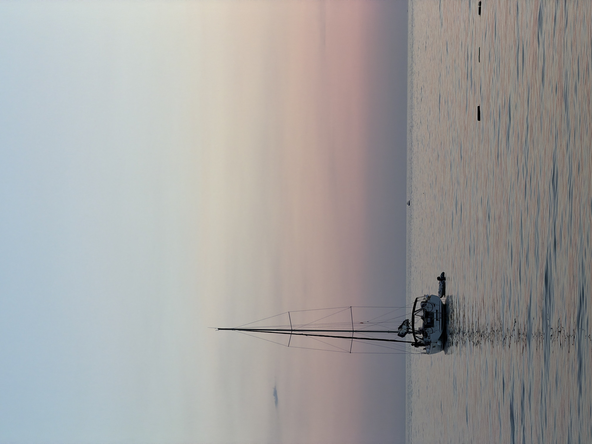 A sailboat is anchored on calm water, its reflection visible below. The sky above is a gradient of pale blue to soft pink, suggesting either dawn or dusk. The sailboat is white with dark trim and has a tall mast with rigging. A small inflatable dinghy is tied to the side of the sailboat. In the distance, there is a very small dark shape that could be another boat. The water has gentle ripples and reflects the colors of the sky. There are also a few dark, rectangular objects visible in the water, possibly buoys or markers. The scene appears serene and peaceful. The location is the sea near Bar, Montenegro.