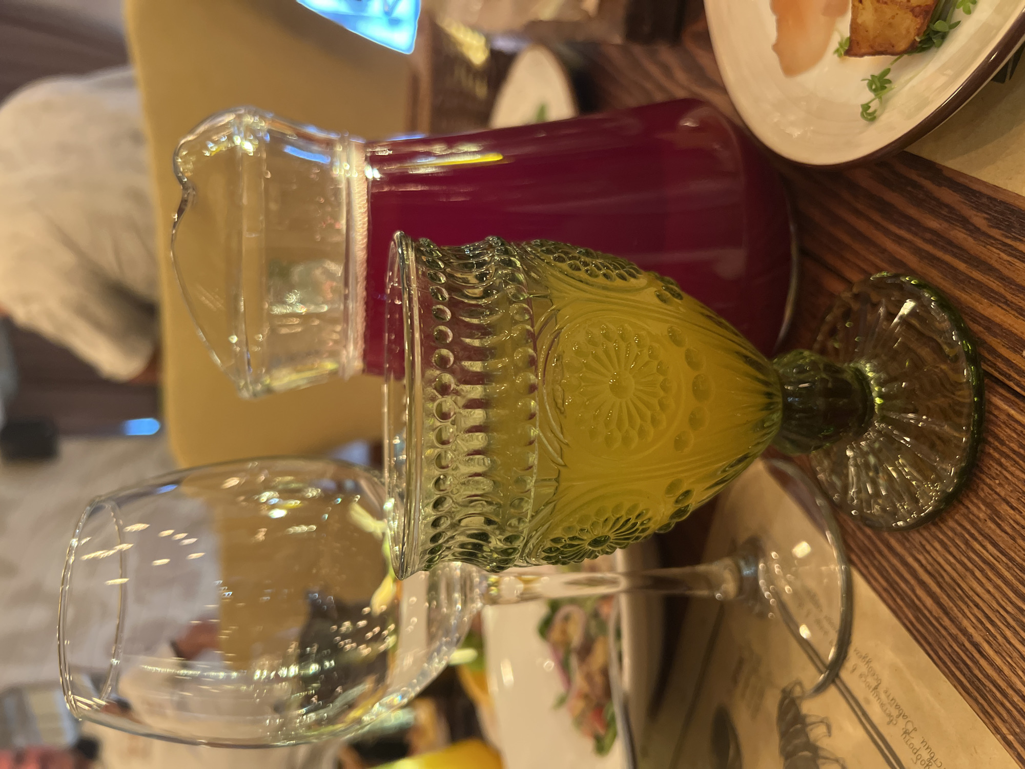 A close-up, eye-level shot showcases a table setting at what appears to be a restaurant in Novosibirsk, Russia. In the foreground, a vintage-style green glass goblet filled with a yellow beverage is prominently displayed. Behind it, a clear glass pitcher holds a deep red liquid. To the left, an empty wine glass stands tall. The table is made of dark wood, and a menu with illustrations is partially visible beneath the wine glass. A white plate with a small portion of food, possibly a slice of fish and a piece of fried potato with microgreens, is on the right side of the frame. In the background, blurry figures of people are seated, and a yellow chair is visible, suggesting a dining environment. The lighting is dim, creating a warm and intimate atmosphere.