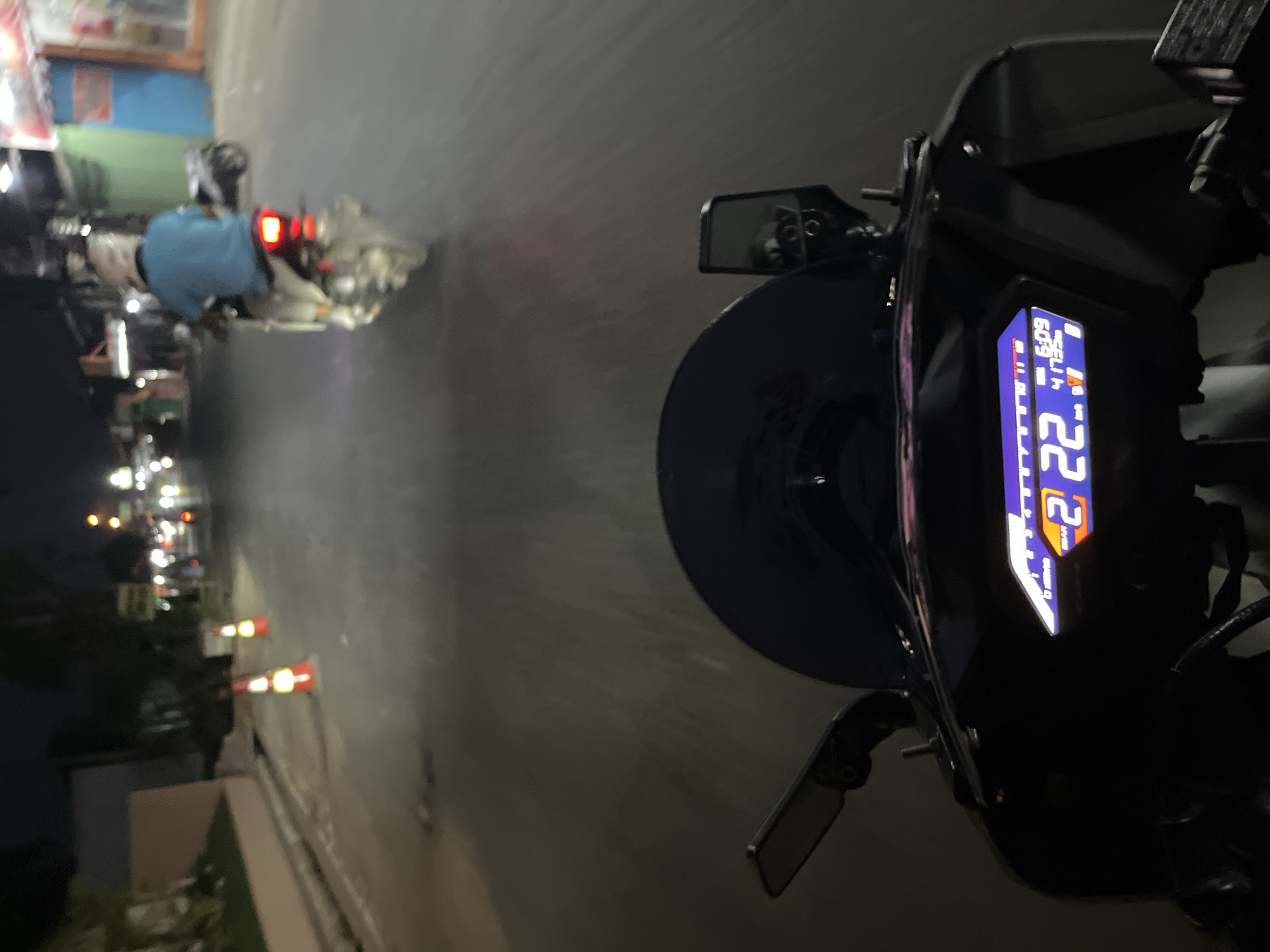 The image captures a nighttime view from the perspective of a rider on a motorcycle in Pecangaan, Indonesia. The road is paved and illuminated by streetlights and the headlights of passing vehicles. In the foreground, the motorcycle's digital dashboard is visible, displaying a speed of 22 km/h, the time 6:09, and an odometer reading of 41733. A dark windshield is positioned above the dashboard. Further ahead, traffic cones are placed on the side of the road, indicating potential road work or a hazard. Another motorcycle with a rider wearing a light blue shirt and a helmet is seen in the middle ground, its taillight illuminated. Buildings with glowing signs line the street on the right side of the image. The overall atmosphere is that of urban nighttime transportation.