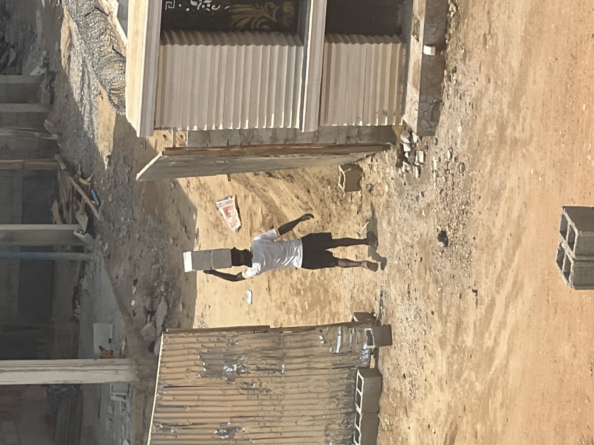 The image depicts an outdoor scene, likely a construction site or an undeveloped area, under bright daylight in Abuja, Nigeria.
The central subject is an adult male, dressed in a white short-sleeved t-shirt, dark shorts, and light-colored shoes. He is actively engaged in manual labor, carrying a grey concrete block (cinder block) balanced on his head, with his right hand extended upwards to steady it.
The setting appears arid and dusty, with the ground composed of bare, light-brown earth and scattered debris, including small rocks and pieces of wood. In the background, there are several structures. To the left, a partially constructed building features a dark facade with elaborate gold-colored patterns, possibly around a doorway or window, and vertical white corrugated siding below. Further left, a dug-out area or trench is visible. On the right side of the frame, another structure is visible, partly covered with corrugated metal sheets, suggesting temporary construction or a work shed. Several unstacked concrete blocks are visible on the ground near these structures.
The bright, direct sunlight indicates it is daytime, casting clear shadows. The overall environment, with its dusty ground, basic construction materials, and the traditional method of carrying heavy loads on the head, aligns with typical scenes in construction or developing areas in Nigeria. There is a red and white plastic bag on the ground near the man, but any text on it is illegible.