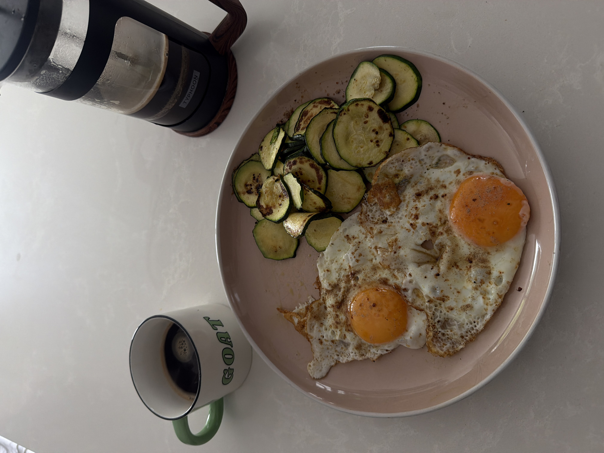 A top-down, slightly angled shot displays a breakfast scene on a light-colored countertop. The main subject is a round, light pink plate filled with two fried eggs and a generous portion of sautéed zucchini slices. The eggs are sunny-side up, with vibrant orange yolks. The zucchini is sliced thinly and appears lightly charred in places, suggesting it has been pan-fried.
To the left of the plate, a white mug with a green handle sits, filled with dark coffee. The mug has the word "GOAT" printed in green lettering. Further left, a French press with a black lid and a glass carafe is visible. The carafe contains some liquid, likely coffee, and a plunger mechanism is visible within. The French press rests on a wooden base.
The lighting suggests daytime, possibly morning given the breakfast context. The overall scene is simple and domestic, implying a casual meal. No people are visible. The background is a neutral, light-colored surface that appears to be a kitchen countertop. There is no indication of weather or specific location other than the general context of a breakfast setting. The visible text on the French press is "TYPHOON" and on the mug is "GOAT".