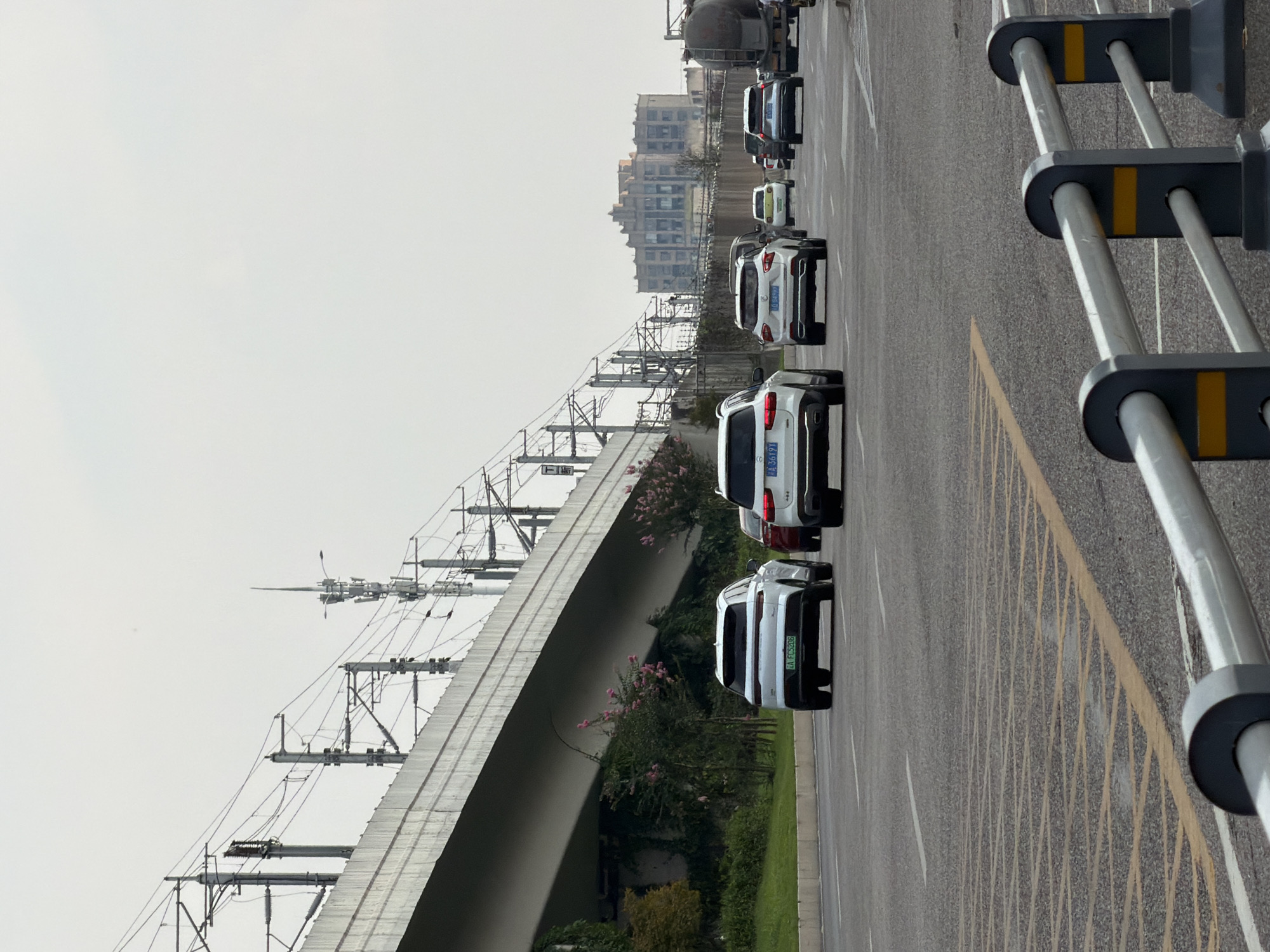 This image depicts a street scene in Jiangjin District, Chongqing City, China. The foreground features a grey asphalt road with yellow and white lane markings. A white metal barrier with black posts and yellow accents runs along the bottom right of the image. In the middle ground, several cars are driving on the road. The cars include a white SUV with license plate "EA FCS208" in the left lane, another white SUV behind it, a red car, and other vehicles further down the road. In the background, a large elevated concrete structure, likely an overpass or bridge, dominates the left side of the frame. Underneath the overpass, there is lush green vegetation with some pink flowering bushes. Electrical poles and wires are visible above the overpass, suggesting a transportation artery or power lines. Further in the distance, on the right side of the image, there are multi-story buildings and a large industrial-looking structure, possibly a factory or storage facility, with a tanker truck visible. The sky is overcast and pale.
