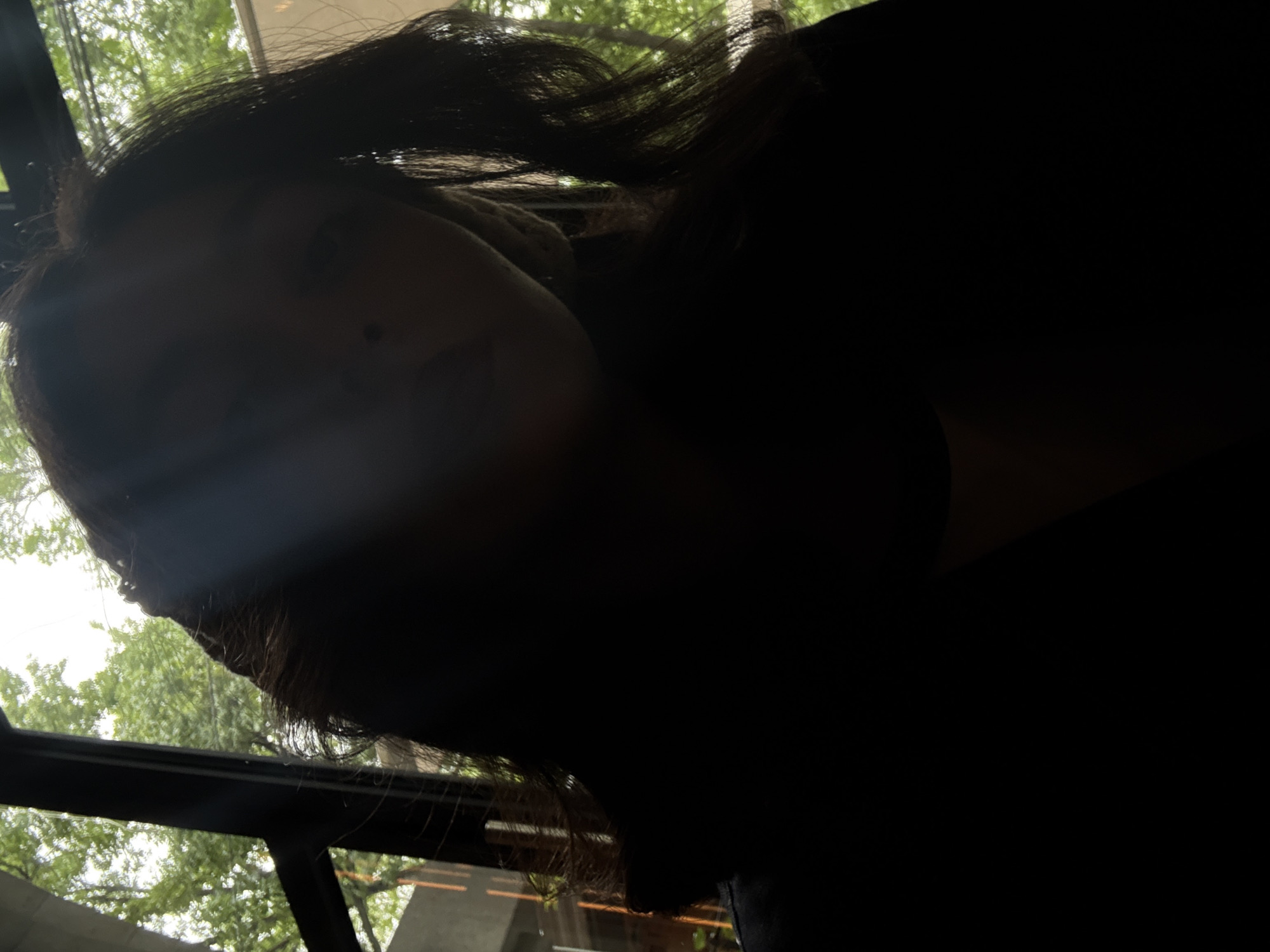 An indoor, low-angle shot captures a person, likely a woman, in a striking silhouette against a brightly lit outdoor background. The subject, positioned in the foreground and viewed from below, has dark, wavy hair cascading around her head. Her face is predominantly in shadow, but faint outlines of her forehead, nose, mouth, and chin are discernible, with a light streak crossing her face. A small dark mole is visible on her right cheek. She is wearing dark clothing that blends into the heavy shadows covering the majority of her body. Behind her head, a light-colored, textured object, possibly a scarf or cushion, contrasts with the dark tones.
The background reveals a vibrant outdoor scene through large windows or a glass roof. Lush green trees and foliage are prominently visible against a bright, overcast sky, suggesting daytime. The dark frames of the windows or structural elements of the building are also apparent. The strong backlighting creates a dramatic contrast, emphasizing the person's form against the luminous exterior. The person appears to be looking upwards, possibly gazing out at the view. There is no visible text. The setting implies a comfortable indoor space with expansive views of nature, potentially a cafe or conservatory.