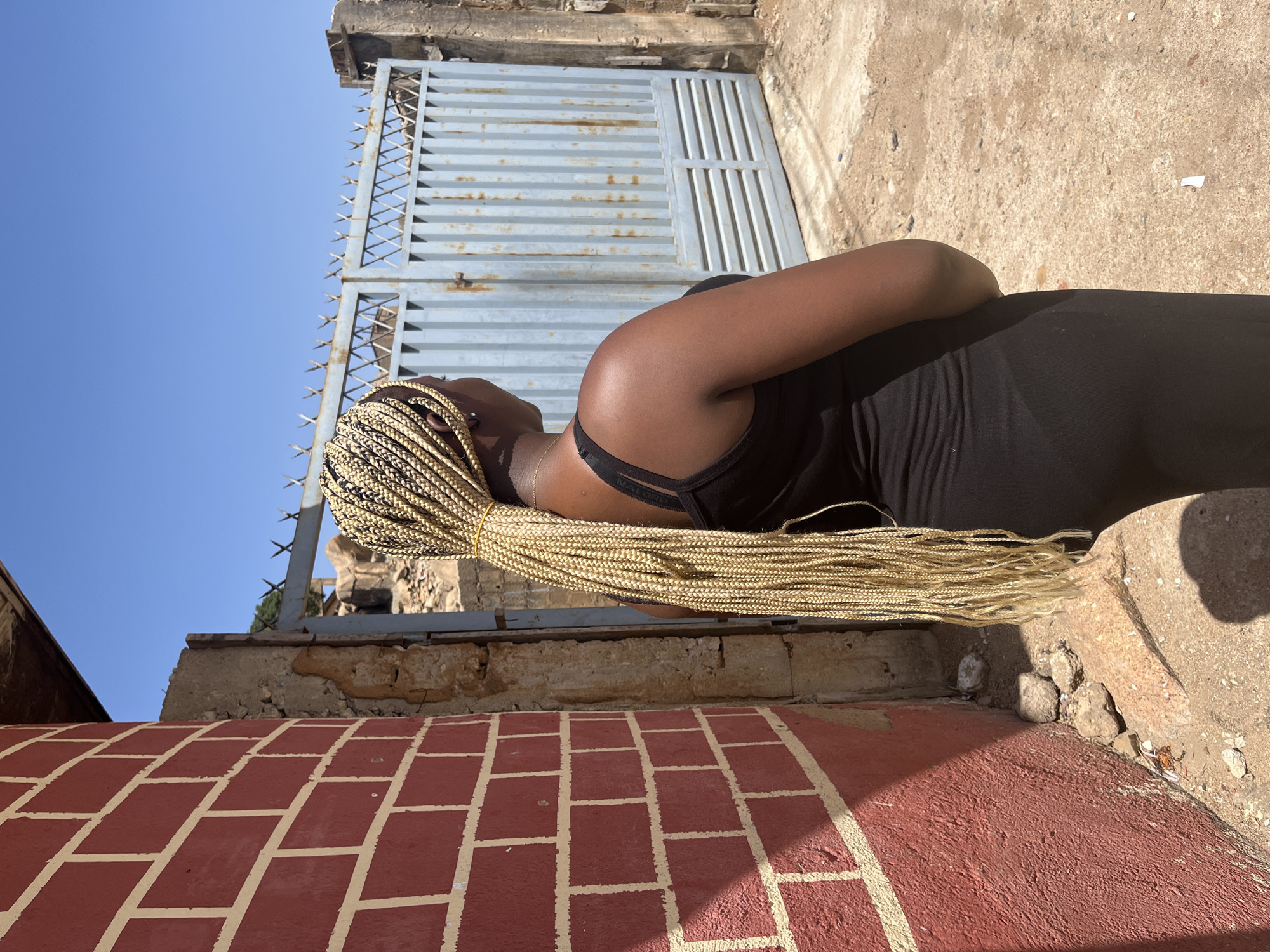 A medium shot depicts a Black woman in Unknown, Unknown, with braided blonde hair, facing away from the camera, in front of a gray gate. She is wearing a black tank top. Her arms are crossed.
The gate, constructed from metal with a spike-topped design, stands slightly ajar, revealing a clear blue sky in the background. The brick structure beneath the gate has faded red bricks and aging material. To the right, a weathered, rough concrete surface extends.