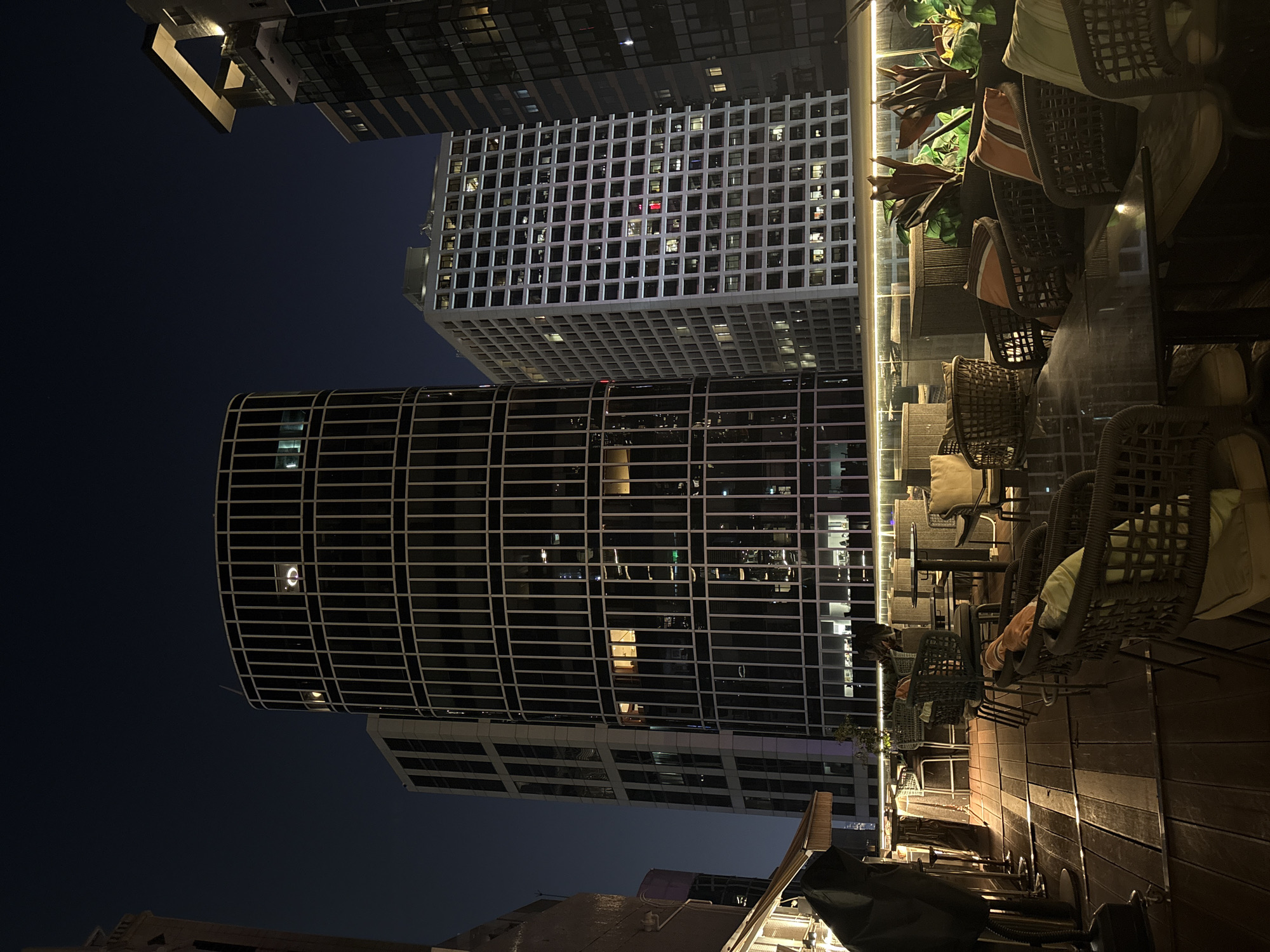 The image captures a rooftop bar or restaurant scene in Hong Kong at night. The foreground features tables and chairs arranged on a wooden deck, typical of outdoor dining settings. The backdrop showcases Hong Kong's distinctive skyline. Two prominent buildings are visible: the Lippo Centre, a cylindrical structure with horizontal bands of windows, and the Hong Kong Arts Centre, known for its grid-like facade. The dark sky suggests a late evening or night setting.