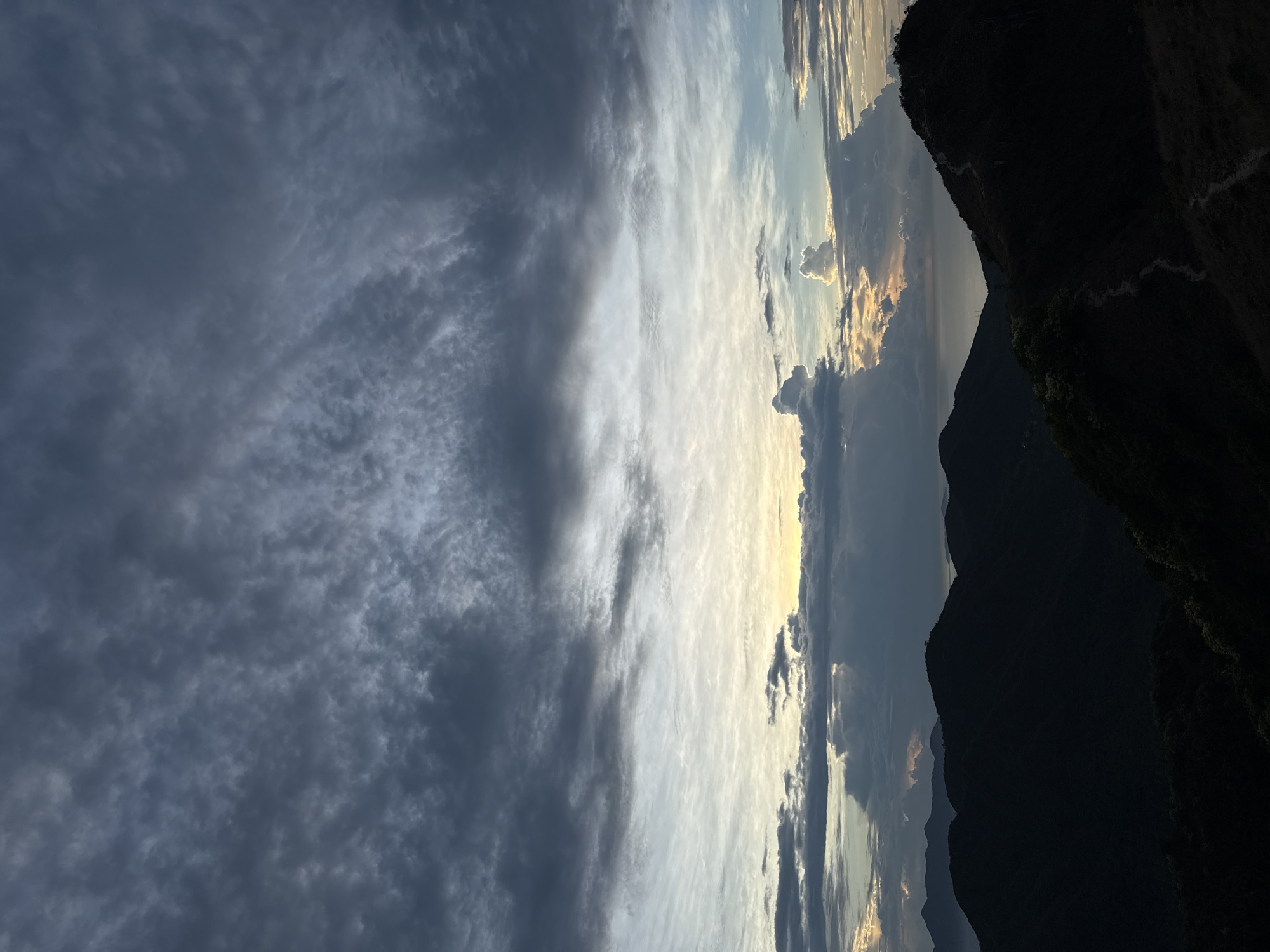 The image presents a landscape view with prominent cloud formations and mountainous terrain. The sky occupies a significant portion of the frame, displaying a mix of dark, heavy clouds on the left transitioning to lighter, brighter clouds near the center and right. Hints of sunset coloration, including golden and orange hues, are visible through gaps in the clouds.
The right side of the image features silhouetted mountain peaks, indicating a rugged, natural environment. A faint, winding trail can be discerned on the side of one of the mountains.
The image suggests a location within a mountainous region, possibly at dusk or dawn, and while the specific ward in Bac Nha Trang is not definitively identifiable within the image, the terrain is consistent with the general geography of the area.