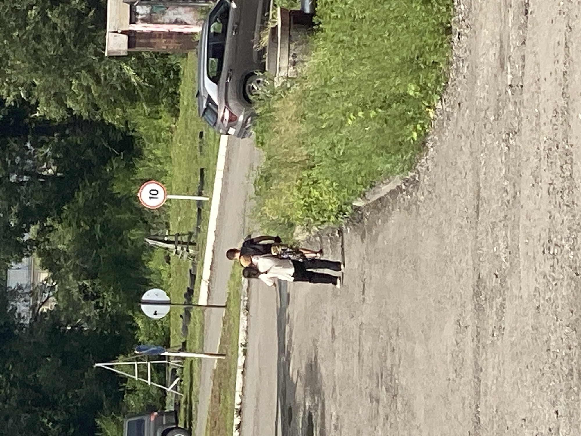 In Novokuznetsk, Russia, two individuals, a person in a white shirt and black pants and another person in a dark shirt, walk away from the camera on a paved path. Behind them, a speed limit sign shows "10" within a red circle. A dark-colored car is parked on the right side of the image, and in the background, lush green trees and bushes dominate the landscape. A ladder-like structure stands to the left, and a small, weathered building is visible on the far right.