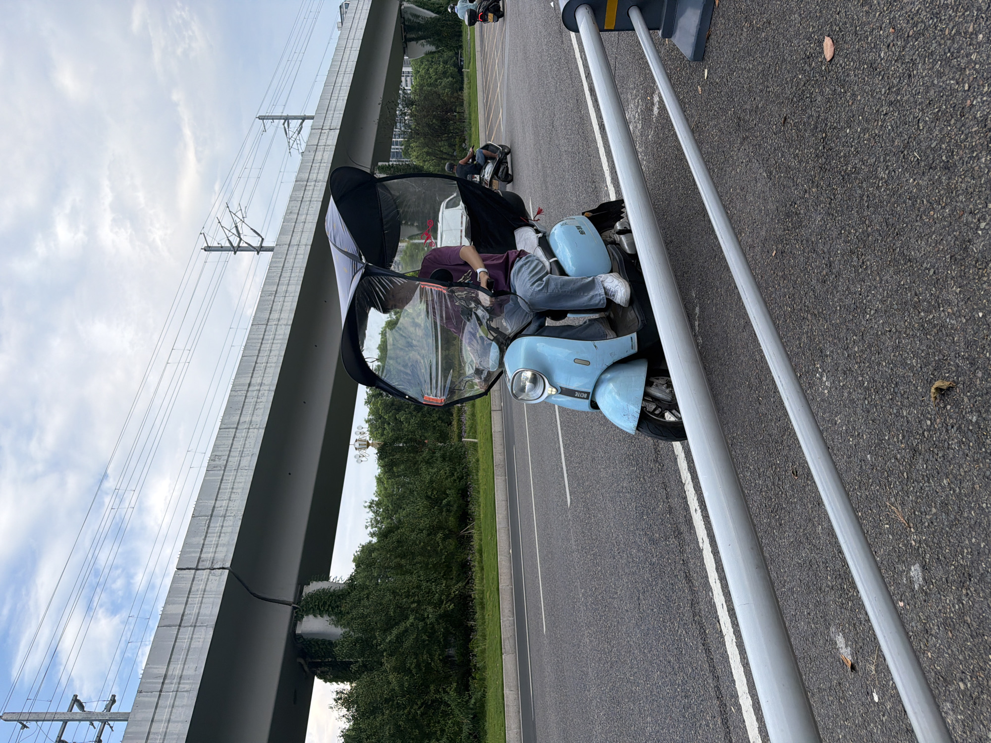 A person is riding a light blue scooter equipped with a transparent canopy and a dark umbrella-like roof in the city of Jiangjin, China. The scooter is on an asphalt road with white lane markings. In the background, there is an elevated concrete structure with overhead electrical wires, indicative of a railway or highway overpass. Lush green trees and vegetation line the side of the road, and a glimpse of buildings can be seen further in the distance. The sky is overcast with scattered clouds. Other scooters are visible further down the road. The scene appears to be during daylight hours.