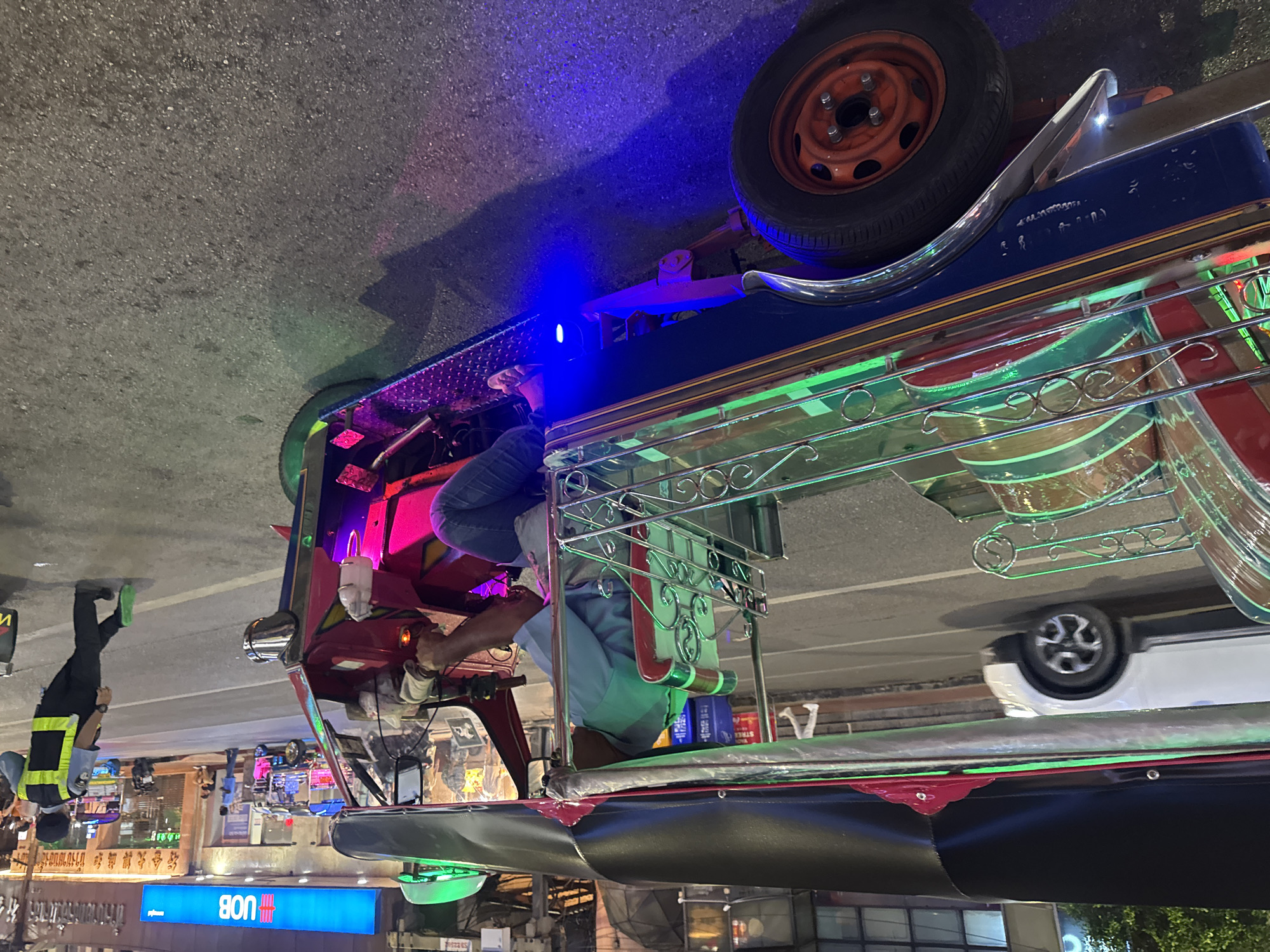 The image captures a tuk-tuk driver and a passenger on the streets of Bangkok, Thailand. The tuk-tuk, a three-wheeled motorized vehicle, is brightly illuminated with neon lights of green, pink, and blue. A spare tire is visible atop the tuk-tuk. The driver is dark-skinned, wearing a light-colored shirt, and is operating the vehicle. A passenger, wearing jeans and a light-colored shirt, is seated behind the driver. A traffic officer in a yellow safety vest stands nearby with light-colored socks and dark shoes. A white car is partially visible behind the tuk-tuk. The street is illuminated by neon signs, one of which reads "UOB" in blue. Buildings stand in the background, and the street appears to be wet or shiny.