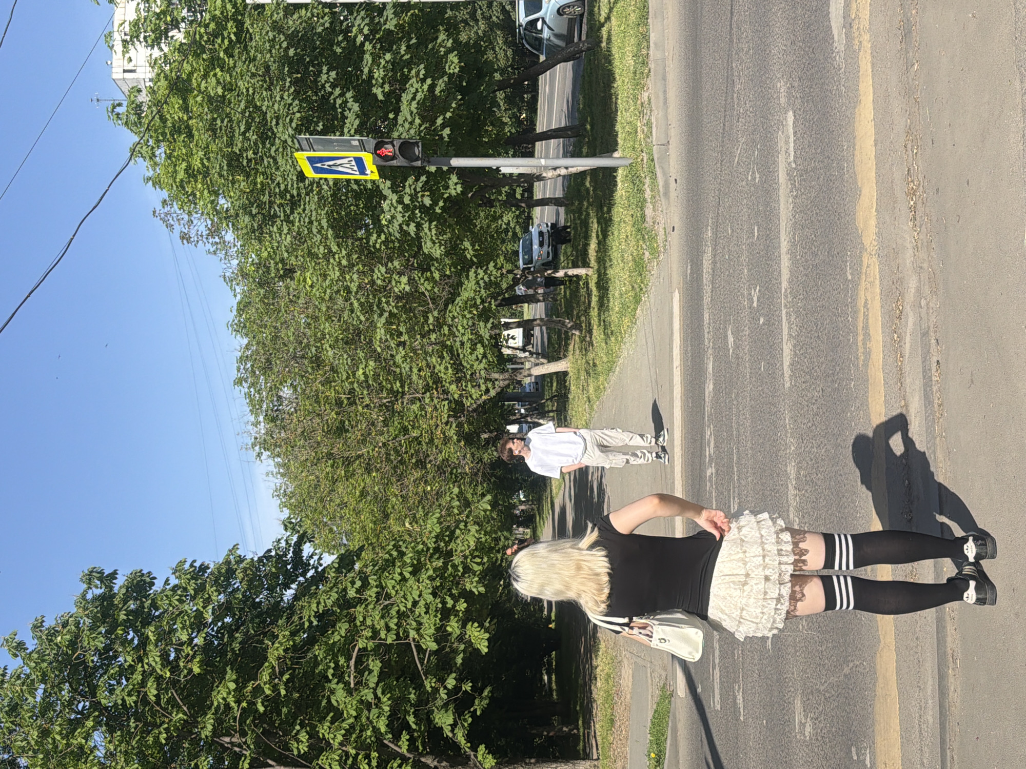 A young woman with long blonde hair is walking on a sidewalk in Rostov-on-Don, Russia. She is wearing a black shirt, a white lace skirt, black and white striped knee-high socks, and black shoes. She also has a white handbag. Another person, wearing a white shirt and tan pants, is walking ahead of her. The sidewalk is adjacent to a street with a yellow line and trees. There is a pedestrian crossing sign with a traffic light nearby. The sky is blue and the day is sunny.