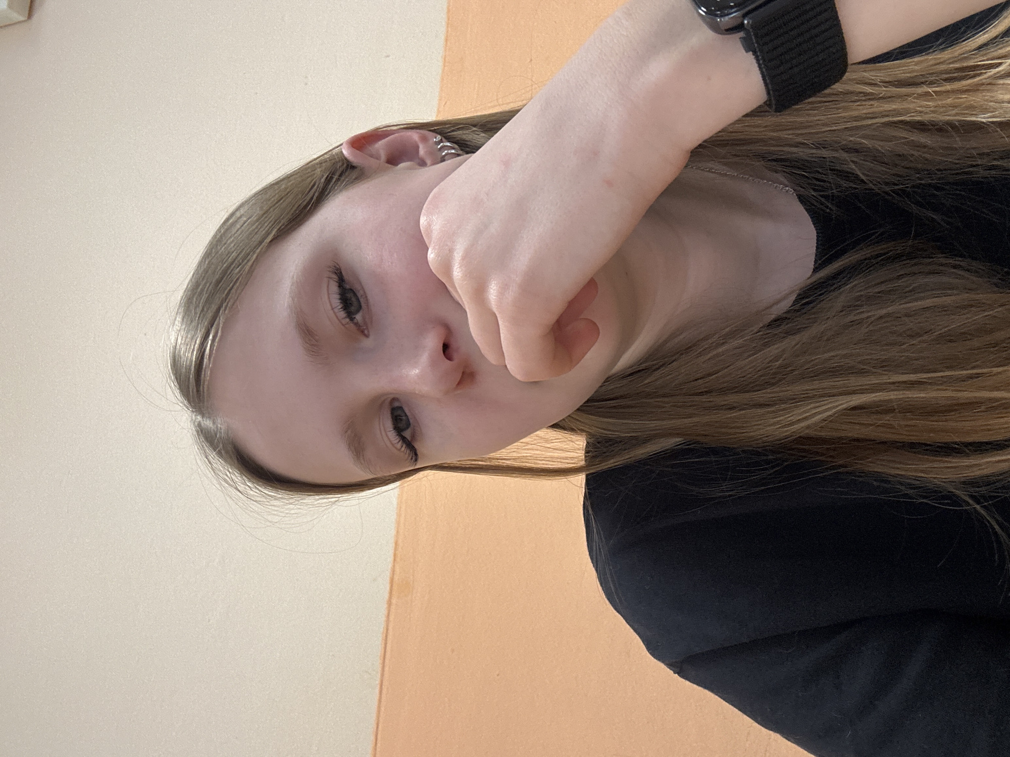 A young woman with light brown hair, wearing a black shirt, is positioned against a two-toned background. The upper left section of the background is a light beige color, while the lower right is a muted orange. The woman's face is partially obscured by her hand, which is covering her mouth. A digital watch is visible on her wrist. There is no indication of the location being Chaikovsky, Russia.