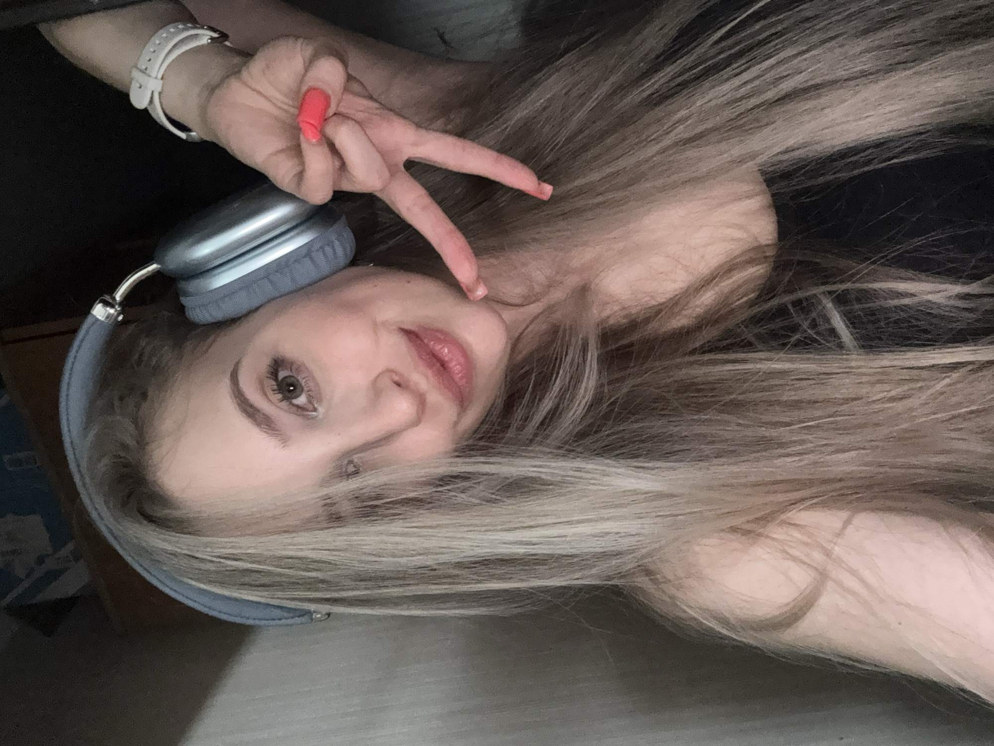 This media file is a close-up, selfie-style image of a young woman lying down. She has long, blonde hair that is spread out around her. She is wearing light blue over-ear headphones. Her left hand is raised, with a coral-pink nail polish on her fingernails, forming a peace sign gesture towards the camera. A white watch is visible on her left wrist. Her face is partially visible, with her lips slightly parted and her eyes looking towards the camera. The overall mood seems casual and relaxed. The lighting suggests an indoor setting, possibly at night or in a dimly lit room. There is no visible text in the image. The location context is Krasnoyarsk, Russia.