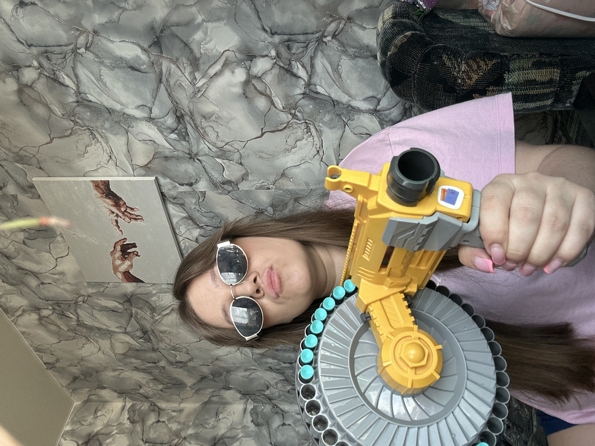 A young person is lying down holding a toy gun, which appears to be a Nerf blaster, towards the camera. The toy is yellow, grey, and blue. They are wearing sunglasses and a pink shirt. A painting that seems to be a depiction of Michelangelo's "The Creation of Adam" is visible in the background. The background has a marble-like pattern. The setting appears to be indoors. It is unclear if the location is in Ольховка, Russia, as there is no information available to verify this.