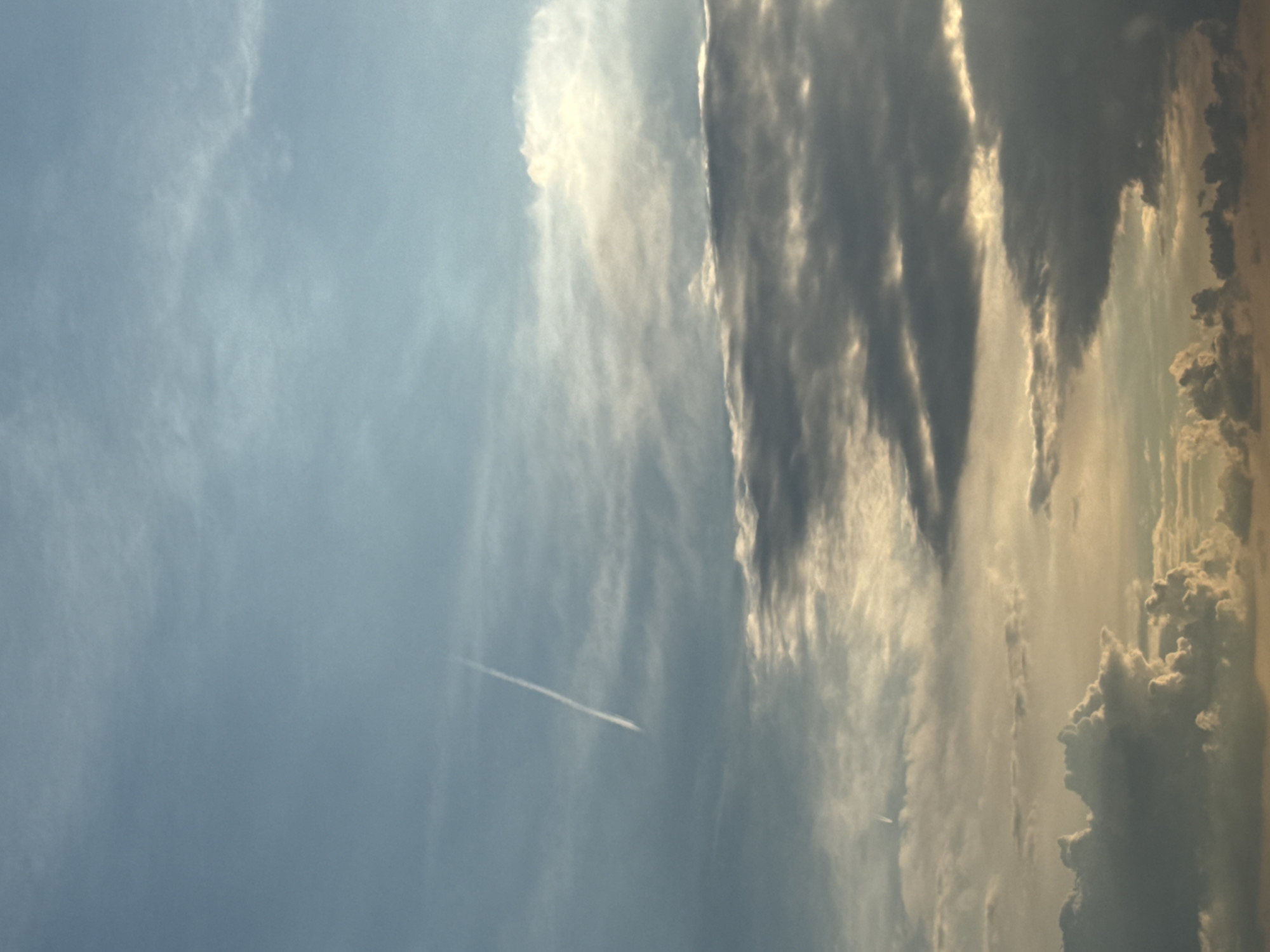 The image shows a daytime sky in Lyubertsy, Russia. There are clouds of various sizes and formations, ranging from wispy cirrus clouds to larger, cumulonimbus clouds with distinct edges. A contrail from an airplane is visible in the lighter blue section of the sky.