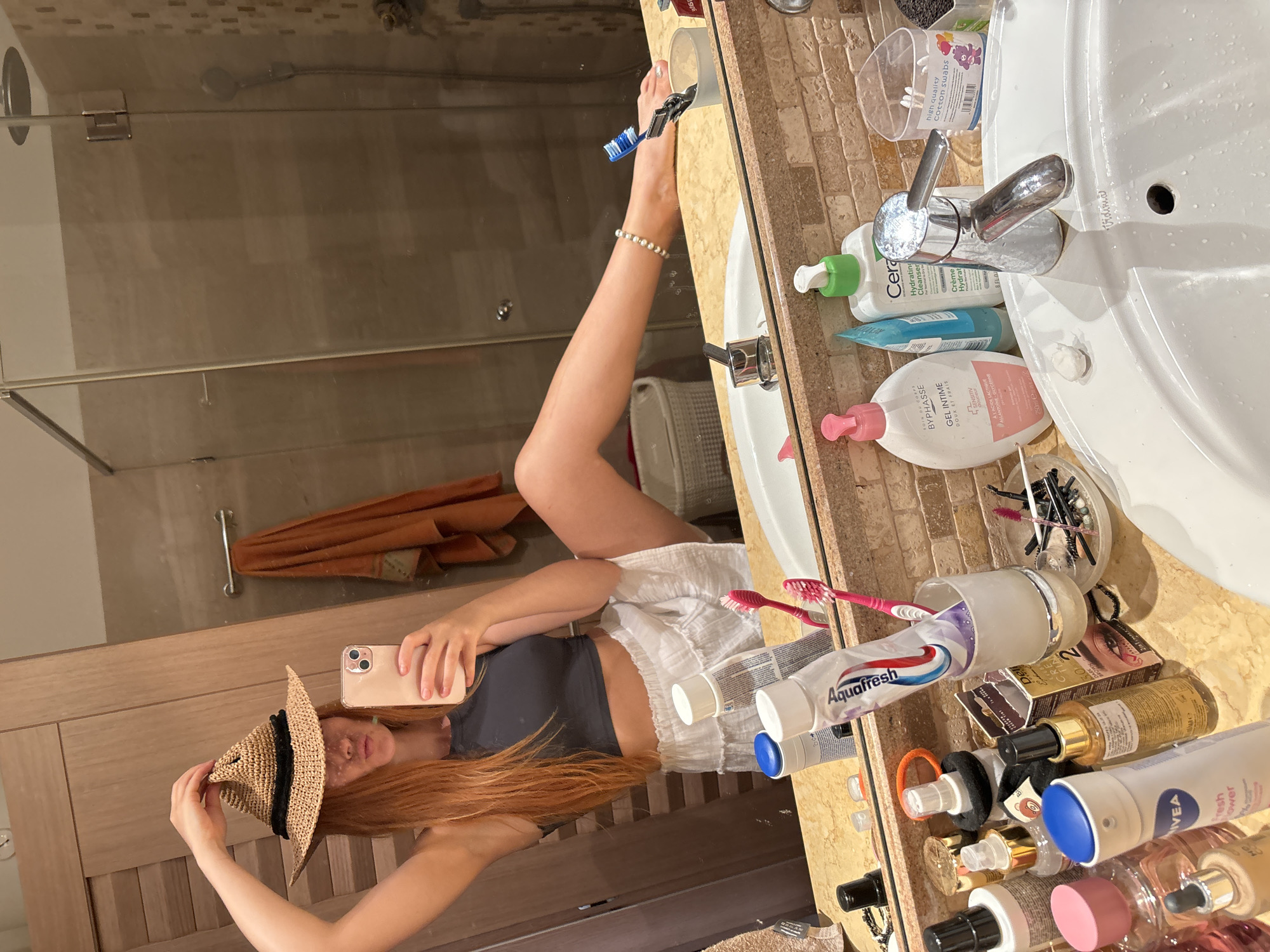 A young woman with long, reddish-blonde hair is taking a mirror selfie in a bathroom. She is wearing a dark gray tank top and white shorts, and has a straw hat tilted on her head. Her right leg is extended upwards, resting on the edge of the sink counter, with a toothbrush held between her toes. The bathroom counter is made of beige tiled material and is cluttered with various toiletries, including bottles of cleanser and lotion, tubes of toothpaste, and makeup brushes. A chrome faucet is visible above the sink. In the background, a glass shower door and a towel rack with an orange towel are discernible. The lighting suggests it is either daytime or well-lit artificial lighting. The image captures a playful and casual moment. Visible text includes brand names like "Aquafresh," "CeraVe," "BYPHASSE," and "NIVEA," as well as "high quality Cotton swabs." The location is identified as Budva, Montenegro.
