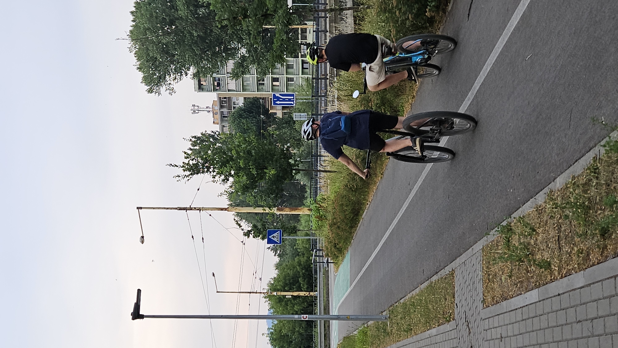 Two individuals on bicycles ride along a paved path bordered by grass and trees. The person in the foreground wears a dark blue t-shirt, black shorts, a white helmet, and a light blue backpack. They are on a bicycle with black handlebars and wheels. The person behind them wears a black t-shirt, light-colored shorts, and a yellow helmet. Their bicycle is blue with black wheels.
The scene appears to be outdoors in an urban or suburban area, with buildings and utility poles visible in the background. A traffic sign indicating a pedestrian crossing is visible. The sky is overcast, suggesting it is likely daytime, possibly late afternoon or early evening. The overall mood seems casual and active, with the individuals enjoying a bike ride. The location is identified as Prešov, Slovakia. No visible text other than signage.