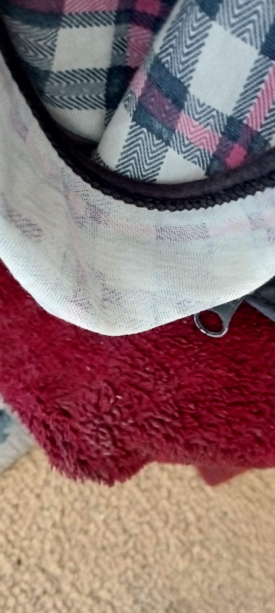 The image shows a close-up, abstract view of textured fabrics. A piece of white fabric with a dark piped edge and a black and pink plaid pattern is draped over a deep red, plush, fuzzy material. The plaid pattern features a herringbone weave within the squares. Below the red fabric, a beige, textured surface, possibly carpet or upholstery, is visible. The lighting is diffuse, and the composition is dominated by the contrasting textures and colors of the textiles. There are no people or discernible landmarks indicating a specific location within Sokuluk, Kyrgyzstan. The image appears to be a still life focused on fabric textures and patterns.
