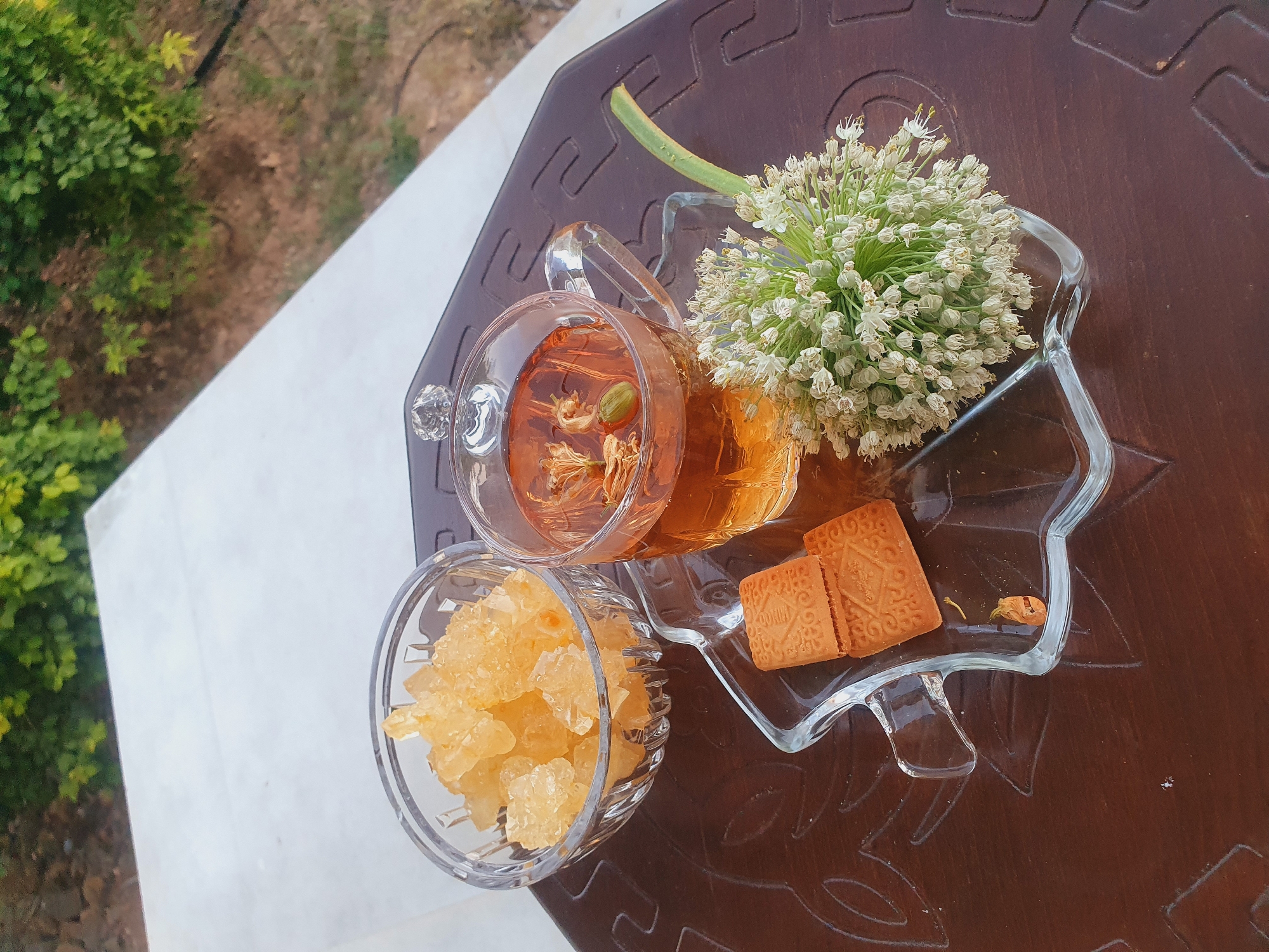 A high-angle, top-down shot captures a tea set arranged on a dark brown, textured table. The tea set includes a clear glass teapot filled with amber-colored liquid, within which are visible cardamom pods and what appear to be dried flower petals. Next to the teapot is a small, clear glass bowl filled with light yellow, crystalline sugar chunks. Two square biscuits, each embossed with a decorative pattern, are placed on a star-shaped, clear glass dish. Beside the biscuits, a small dried flower petal rests on the dish. A large cluster of small white flowers on a green stem is also placed on the star-shaped dish, adding a decorative element.
The background suggests an outdoor setting, with lush green foliage visible on the left and a blurred earthy area beyond. The lighting indicates it is daytime, likely late morning or early afternoon, with soft natural light. The overall scene evokes a sense of relaxation and a pleasant moment of enjoying tea. No visible text is present in the image. The location context of Kheyrabad, Iran, suggests a potentially warm climate.
