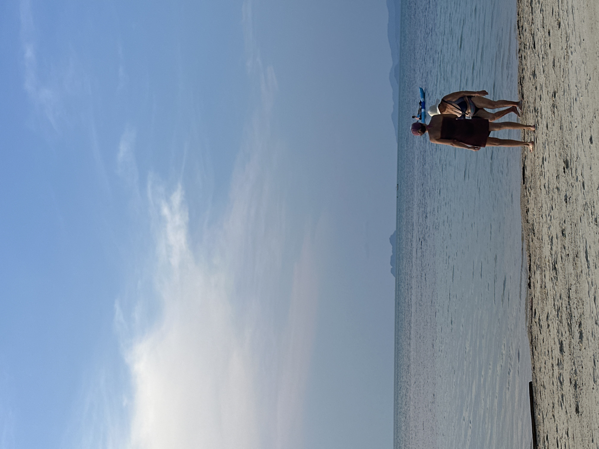 This full-shot landscape image captures a peaceful beach scene at Baan Tai, Thailand, under a clear, bright blue sky with scattered wispy clouds. The horizon line is visible, separating the sky from a calm, slightly rippled body of water, likely the sea.
In the foreground, a strip of sandy beach runs horizontally across the lower part of the frame. Two individuals are standing on the sand, facing the water. The person closer to the viewer is seen from behind, wearing a dark reddish-brown wrap or towel and a head covering of a similar color. Slightly behind and to their right stands another person, also seen from behind, wearing a striped swimsuit and a light-colored hat. This individual is holding a blue object, possibly a small surfboard or snorkeling gear, indicating engagement in or preparation for water activities.
In the mid-ground, the expansive, tranquil water stretches out towards distant, hazy landmasses or mountains visible on the horizon. The lighting suggests it is daytime, possibly morning or late afternoon, with soft, even illumination. The overall atmosphere conveyed is one of leisure and calm at a tropical beach destination. There is no visible text in the image.