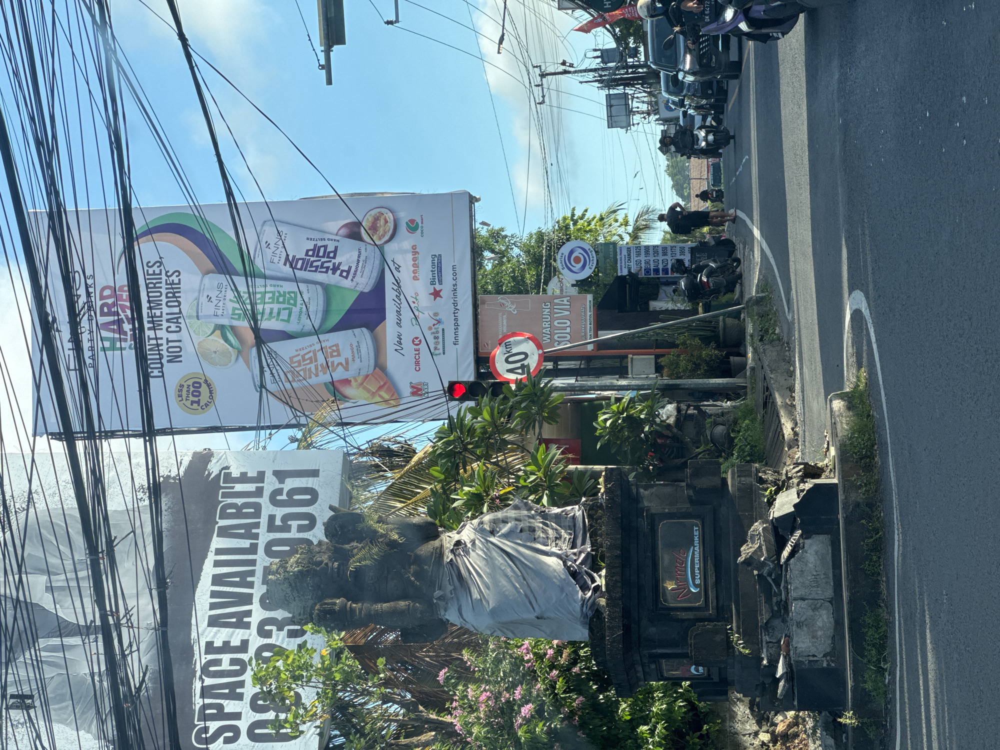 The image shows a street scene in Kuta Selatan, Indonesia. A large billboard advertising "Finns Party Drinks" is prominently displayed above a statue draped in fabric. Below the billboard, another sign advertises "SPACE AVAILABLE" and a phone number. A "40 km" speed limit sign and a sign for "Warung Solo Via" are also visible. A money exchange rates board shows current exchange rates. Several motorcycles are parked along the side of the road. The sky is clear and blue. The "Nirmala Supermarket" sign is visible on the roadside. Overlapping power lines crisscross the sky.