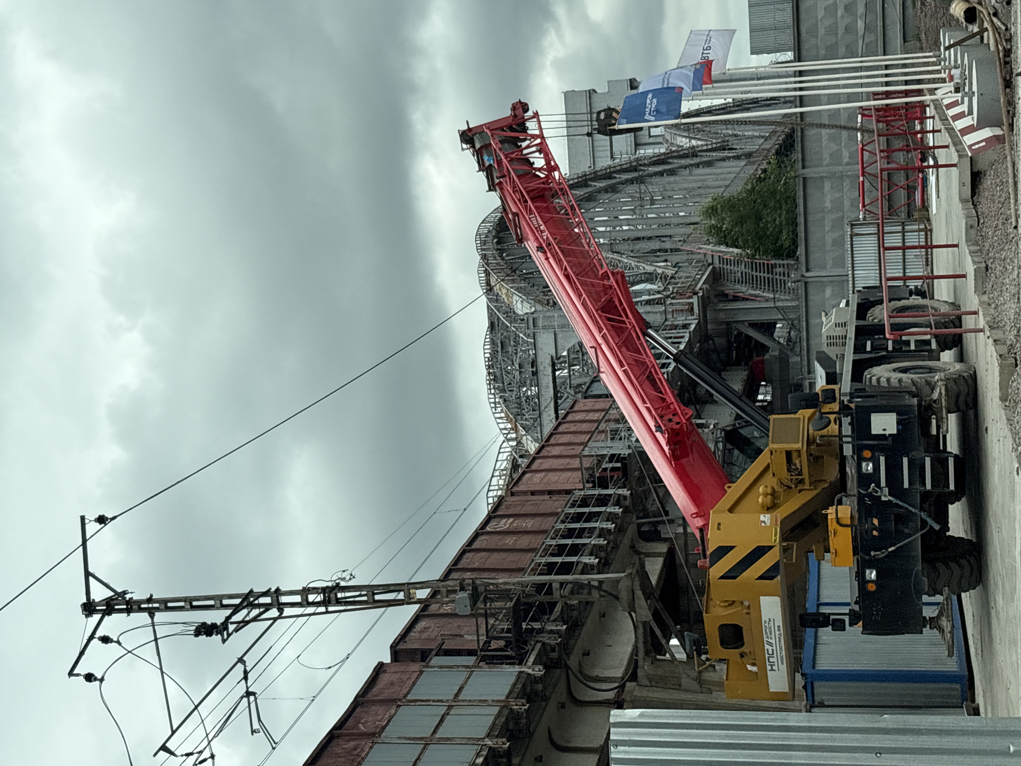 This media file features a large red crane positioned on a construction site under a cloudy sky. The crane's arm extends upwards, dominating the frame. In the background, a large industrial structure with a metal framework is visible, suggesting ongoing construction or renovation. Several flags are displayed on poles, with one clearly showing "ВТБ" (VTB Bank) in large letters, along with another flag with text partially obscured.
The scene appears to be a construction or industrial area in Saint Petersburg, Russia. The lighting suggests it's daytime, but the overcast sky casts a diffused light over the scene, creating a somewhat muted atmosphere. The ground appears to be a mix of gravel and concrete, typical of a construction site.
Details on the crane include its yellow and black counterweight and base, with visible tires and hydraulic components. There is also text on the crane, including "7739" and "PALFINGER". Another piece of text is visible on a white sticker on the crane's base, which appears to read "НПС И МОСТОСТРОЕНИЕ" (NPS and Bridge Construction).
The overall impression is one of industrial activity and heavy machinery at work on a large-scale project. The weather appears to be overcast, contributing to a potentially cool and damp environment. No people are clearly visible in the frame, emphasizing the focus on the machinery and the construction itself.