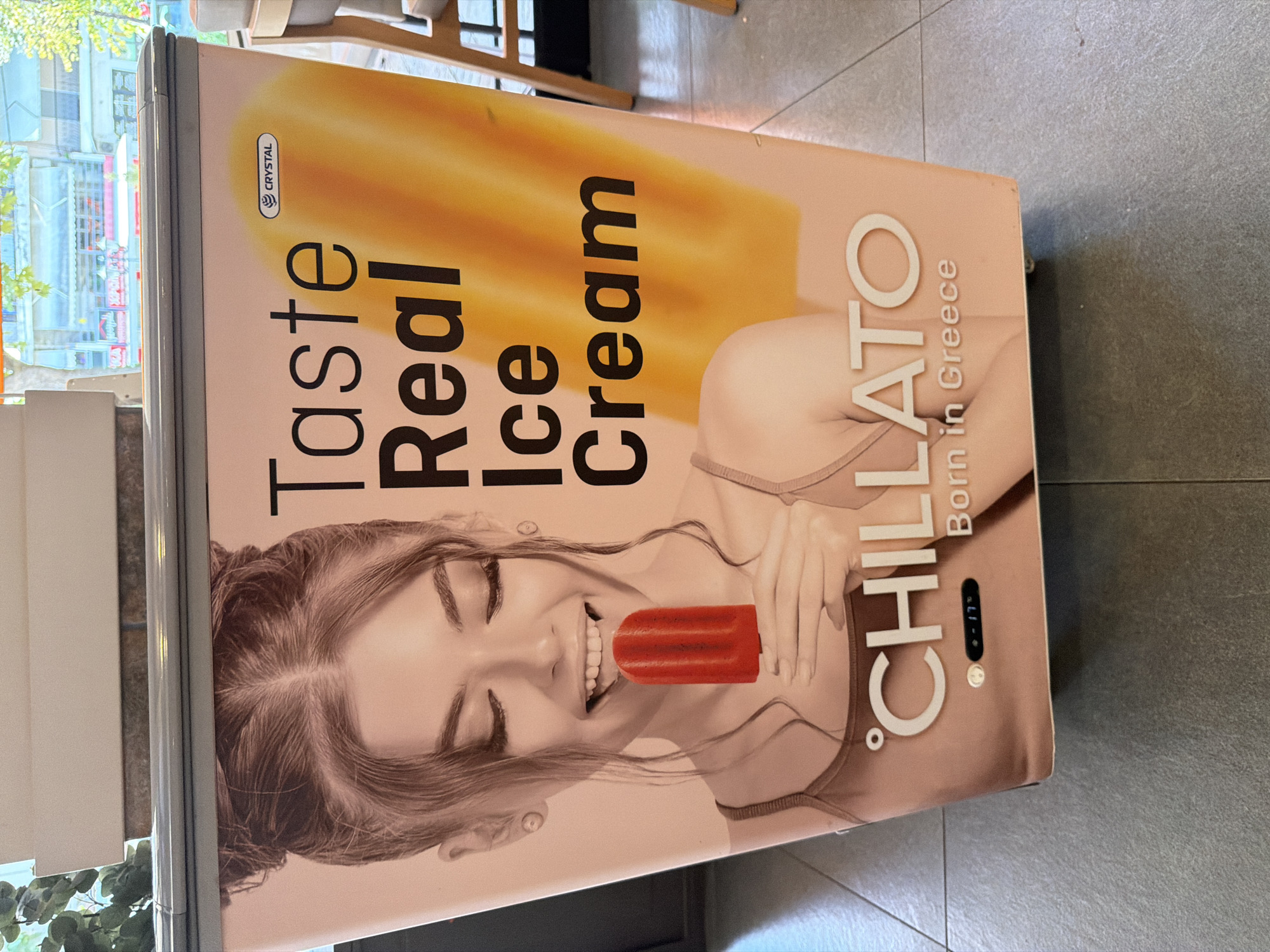 The image shows a freezer advertising "Chillato Born in Greece" ice cream. A young woman with her eyes closed is depicted biting into a red popsicle. The freezer is branded with "Crystal" and displays the text "Taste Real Ice Cream." The freezer is positioned on a grey tiled floor, likely in a shop or establishment, with a blurred background of buildings and trees suggesting an outdoor urban setting in Asprovalta, Greece. A temperature display on the freezer reads "-17°".