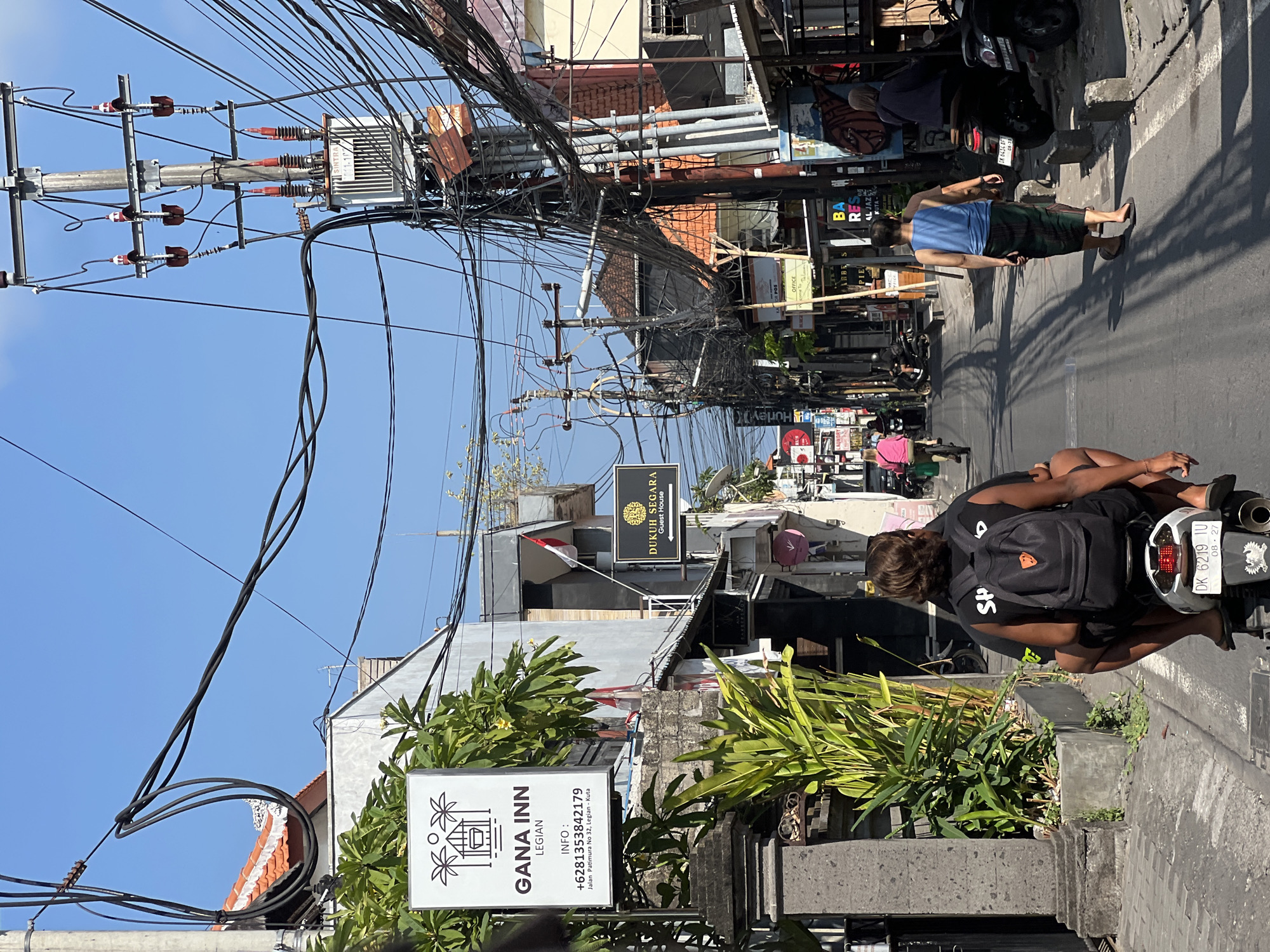 The image depicts a street scene in Kuta, Indonesia, on a bright, sunny day with a clear blue sky. The street is lined with buildings, some of which appear to be commercial establishments, including a sign for "GANA INN LEGIAN" and another for "DUKUM SEGARA Guest House". A prominent feature of the scene is the dense network of electrical wires suspended above the street, running from utility poles. One pole is equipped with a transformer.
In the foreground, two people are riding on a scooter, viewed from behind. The person in front is wearing a black t-shirt with a backpack, and the person behind is wearing a blue tank top and a sarong. Both are wearing sandals. The scooter's license plate reads "DK 6219 IU".
Further down the street, other individuals are visible. A person is seen riding a bicycle in the middle ground. On the right side of the street, two individuals are standing near motorcycles. One is wearing a blue top and a sarong, and another person is partially visible on the right edge of the frame, wearing a dark top.
The environment suggests a bustling urban or tourist area. The architecture of the buildings and the presence of signs in English and Indonesian indicate a location frequented by tourists. The road surface is asphalt, and there are concrete curbs and some foliage planted along the sidewalk. Shadows cast by the buildings and overhead wires are visible on the road, indicating the position of the sun. Several motorcycles are parked along the sides of the street.