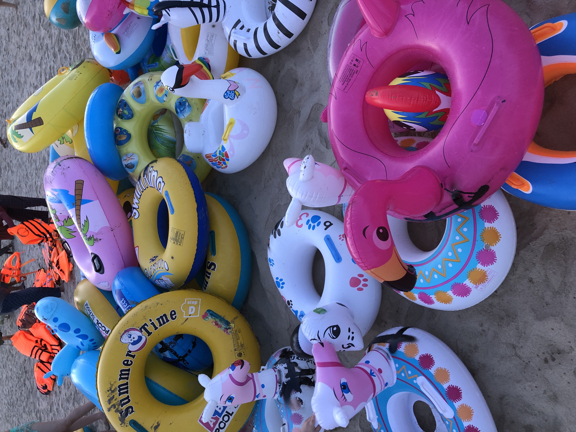 The image depicts a collection of inflatable pool toys and life vests on a sandy beach, likely in Da Nang, Vietnam, given the context. The toys are various shapes and colors, including animal-themed rings, a flamingo ring, and a swan-shaped float. In the background, several orange life vests are piled up, suggesting a rental or recreational area. The sand appears damp, and the overall scene indicates a beachside location associated with water activities. No people are clearly depicted in the immediate foreground, but hints of legs and clothing are visible in the upper left and center-right portions of the image, suggesting a public area.
