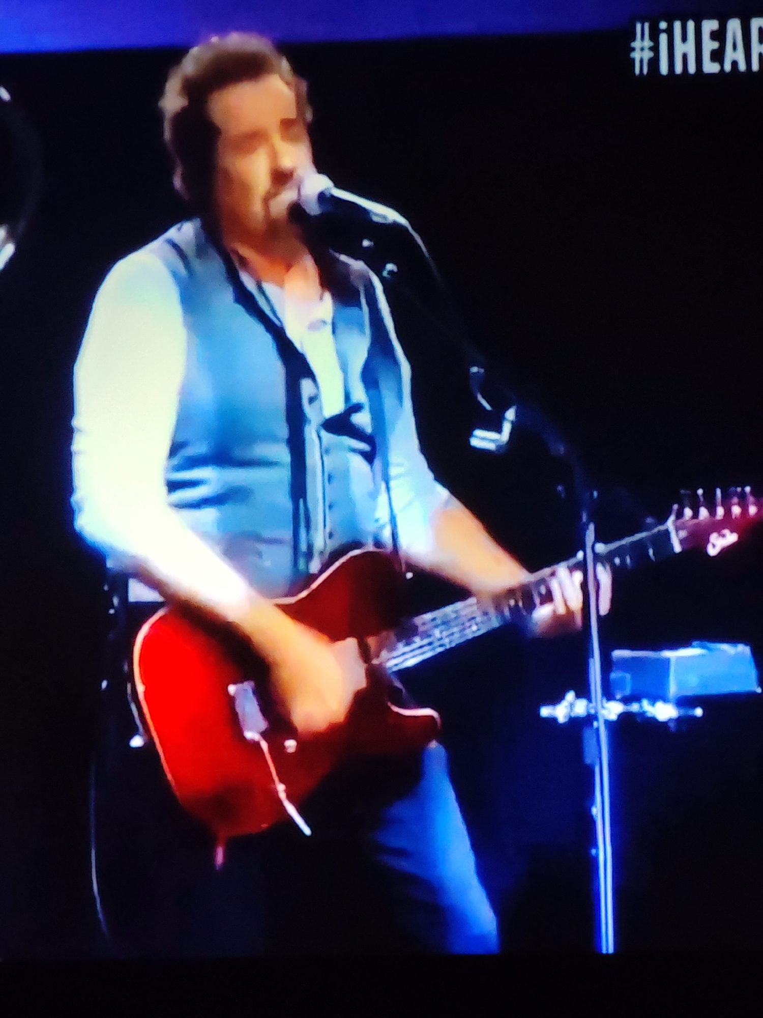 The image shows a man playing a red electric guitar on a dimly lit stage. He is wearing a light blue shirt and a dark blue vest. A microphone is positioned in front of him. In the background, there is a screen displaying the hashtag "#iHEAR" in white text. The overall scene suggests a live musical performance. There is no indication of the location being in Vladimir, Russia.
