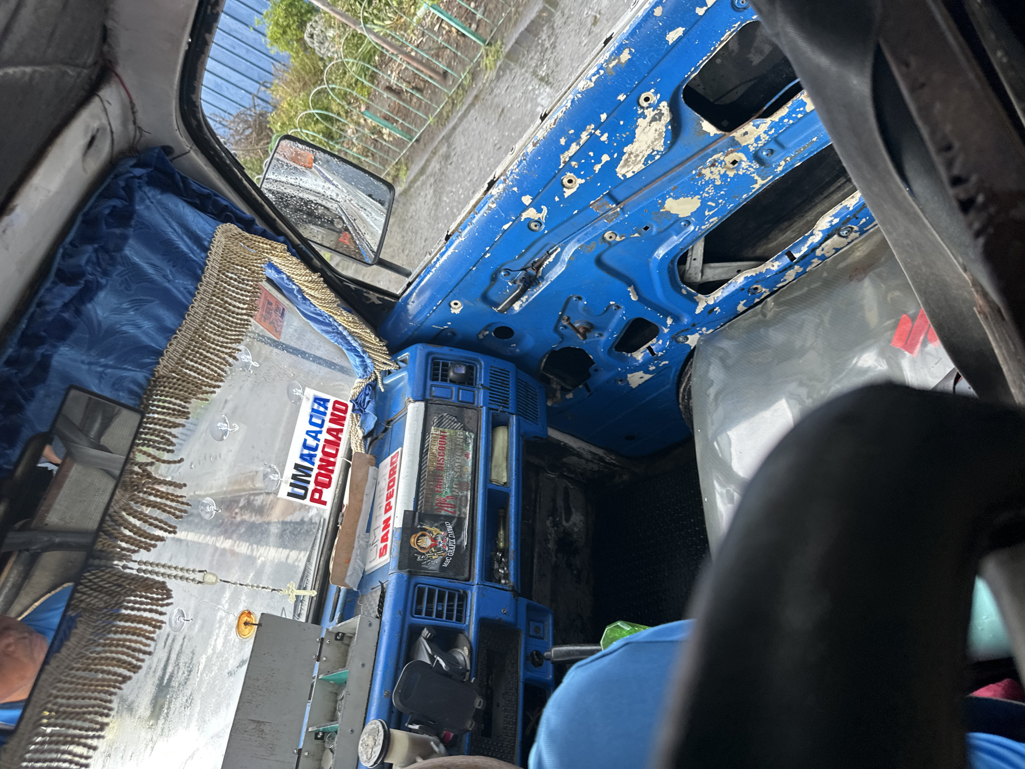The image shows the interior of a jeepney, a type of public transportation common in the Philippines, specifically in Davao City. The dashboard and roof of the vehicle are painted a bright blue. The paint is peeling in several areas, revealing the underlying metal. The windshield is decorated with tassels, stickers that read "UMACACIA PONCIANO", and "SAN PEDRO." There is also a rosary hanging on the windshield. A side mirror is visible in the upper left. The passenger's side of the jeepney appears to be open, exposing the street outside. A person wearing a blue shirt is visible in the lower right of the image, likely a passenger. The overall impression is of an old, heavily customized, and well-used vehicle.