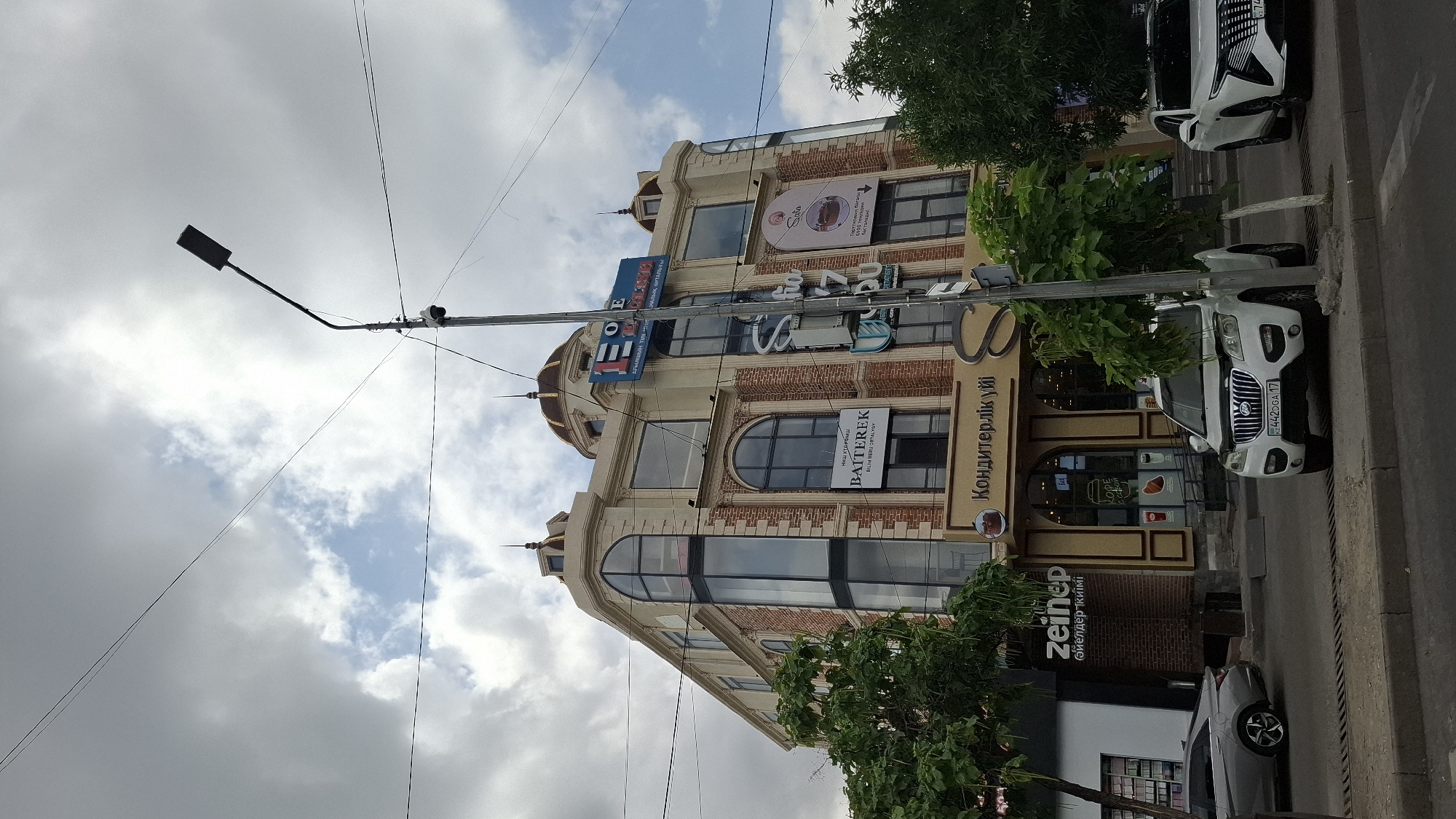 The image shows a modern, multi-story building in Shymkent, Kazakhstan. The building has a light-colored facade with red brick accents and large windows. Several businesses are advertised on the building, including "BATTEREK", "Sofia", and a "Кондитерлік үй" (Confectionery House). A street lamp with a security camera is visible in the foreground, along with two white cars parked on the street. Trees line the sidewalk, and the sky is partly cloudy.