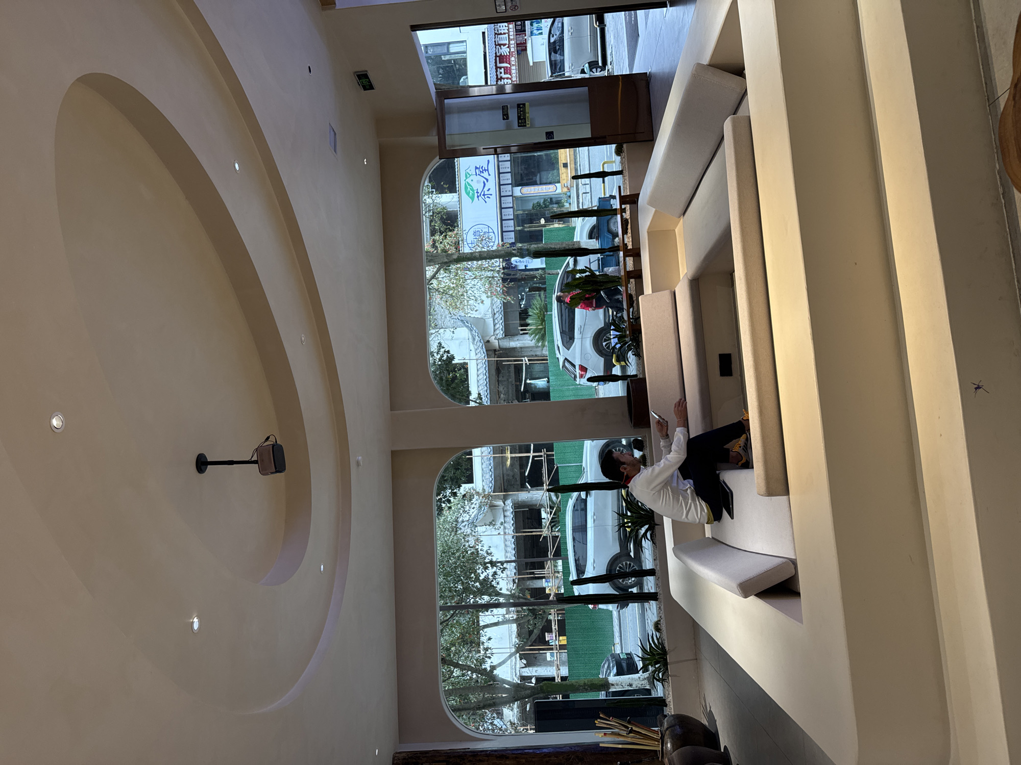 The image displays an interior view of a modern lounge area within a building located in Wulingyuan, China, looking outward onto a city street.
The interior features a light-colored, possibly beige, ceiling with multiple recessed lights and a large, multi-layered circular/oval design. A dark-colored projector hangs from the ceiling. Dominating the foreground and center is a large, light-colored, multi-tiered sunken seating structure with U-shaped light gray cushioned benches and backrests.
One male individual is seated within this sunken lounge, positioned on the inner cushion. He wears a white long-sleeved shirt, dark trousers, and yellow and black athletic shoes. He is actively engaged with a smartphone or small tablet held in his hands, looking down at the device. Another mobile device rests on the seat beside him.
Large, arched windows line the wall, offering a view of the street outside. In the immediate foreground of the window view, a planter containing spiky green succulents and several tall, thin, dark decorative elements is visible. To the far left, two large, dark, textured pots containing bamboo-like sticks are present. To the far right, a wooden door with glass panels and a push bar is visible, with an illuminated "EXIT" sign in both English and Chinese positioned above it.
The street scene outside reveals a paved road, which appears damp. A white SUV is prominently parked or moving slowly in the lane nearest the building. A dark sedan is partially visible to the left of the SUV, and a white van is further down the street to the right. Across the street, low-rise buildings are visible, including one with scaffolding and a green construction barrier, indicating ongoing construction. Another storefront displays Chinese characters, including "茶屋" (Tea House), in blue on a white sign. Trees with sparse foliage line the street. A pedestrian wearing a red jacket or vest is visible walking on the sidewalk across the street.