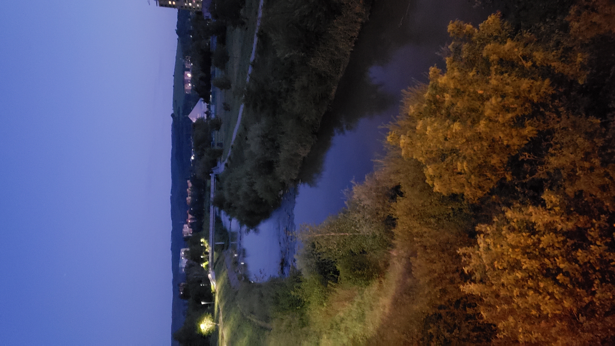 The image shows a view of the Sekčov river in Prešov, Slovakia, at dusk. The river flows through a vegetated area with a walking path along its bank. In the background, there are buildings and a hill with lights visible, indicating a suburban or city area. The sky is a deep blue, typical of twilight. The foreground is dominated by trees and bushes, some of which are illuminated by artificial light, casting an orange glow on their leaves. A bridge crosses the river in the mid-ground.
