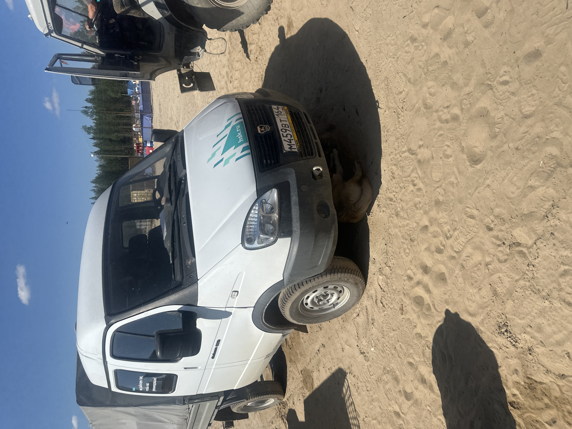 A white van with a gray tarp-covered cargo bed is parked on a sandy surface, likely in Чикшино, Russia. The van has a license plate reading "M459BT 164 RUS". A logo with turquoise geometric shapes and the text "hsk.ru" is visible on the front of the van. A dog is partially visible underneath the front of the van. In the background, a tall, dark-colored vehicle with its door open is parked next to the van. Beyond that, there are trees, tents, and possibly event structures set up on the sandy terrain under a clear blue sky with a few scattered clouds. The ground is covered in sand with visible tire tracks and footprints.