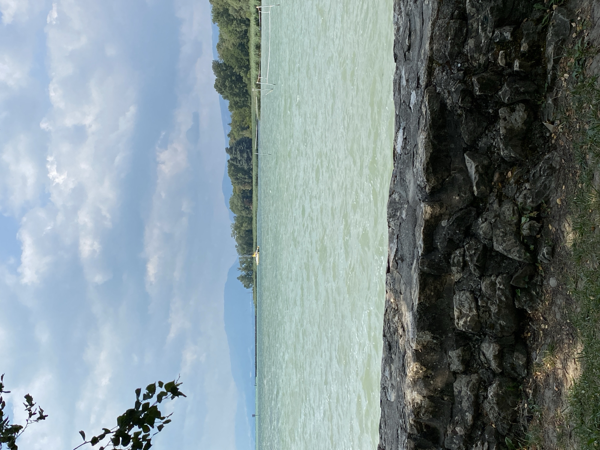 The image shows a body of water with a stone wall in the foreground. Trees and hills are visible in the background. There is a sailboat on the water. The sky is cloudy. The scene appears to be a park or a recreational area by a lake or sea. The location is specified as Laténa, Switzerland.