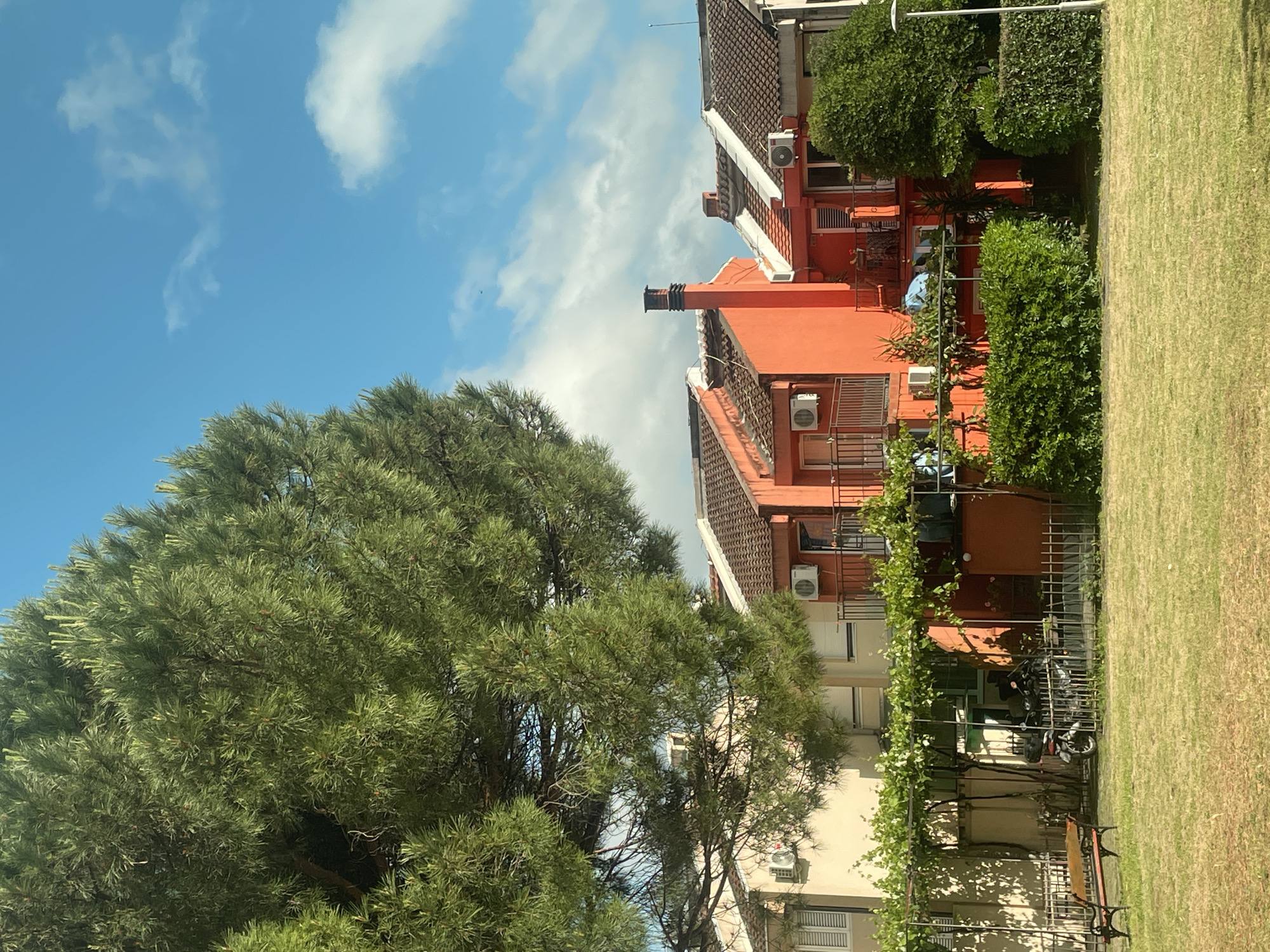 The image depicts a partially obscured view of a residential area. A large, dense tree dominates the left side, concealing the base of a building. To the right of the tree, a row of buildings is visible, some painted orange with reddish-brown tiled roofs. Several air conditioning units are attached to the exterior walls. Between the buildings and the right edge of the frame, there's a patch of grassy land. A fence is present, and what seems to be a motorcycle is parked behind it. A partially visible bench sits nearby. The sky above is blue with scattered white clouds.
