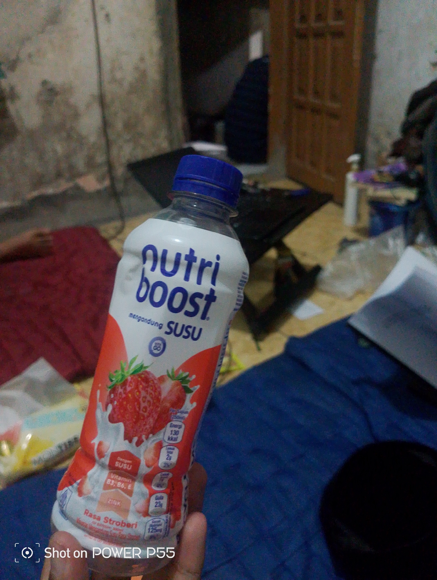 A close-up, eye-level shot features a bottle of NutriBoost strawberry-flavored milk in the foreground, held by a hand on the right. The bottle, with its white label and red strawberry graphics, is tilted slightly. Text on the bottle indicates it contains milk and is enriched with vitamins B3, B6, and E, as well as zinc. The environment appears to be an indoor setting, possibly a room, with various items scattered around. To the left of the bottle, a red fabric is visible, and beyond that, a person's arm is partially obscured. The background is dimly lit, showing indistinct objects like a laptop, a door, and some shelves. The overall impression is of a casual, everyday scene.