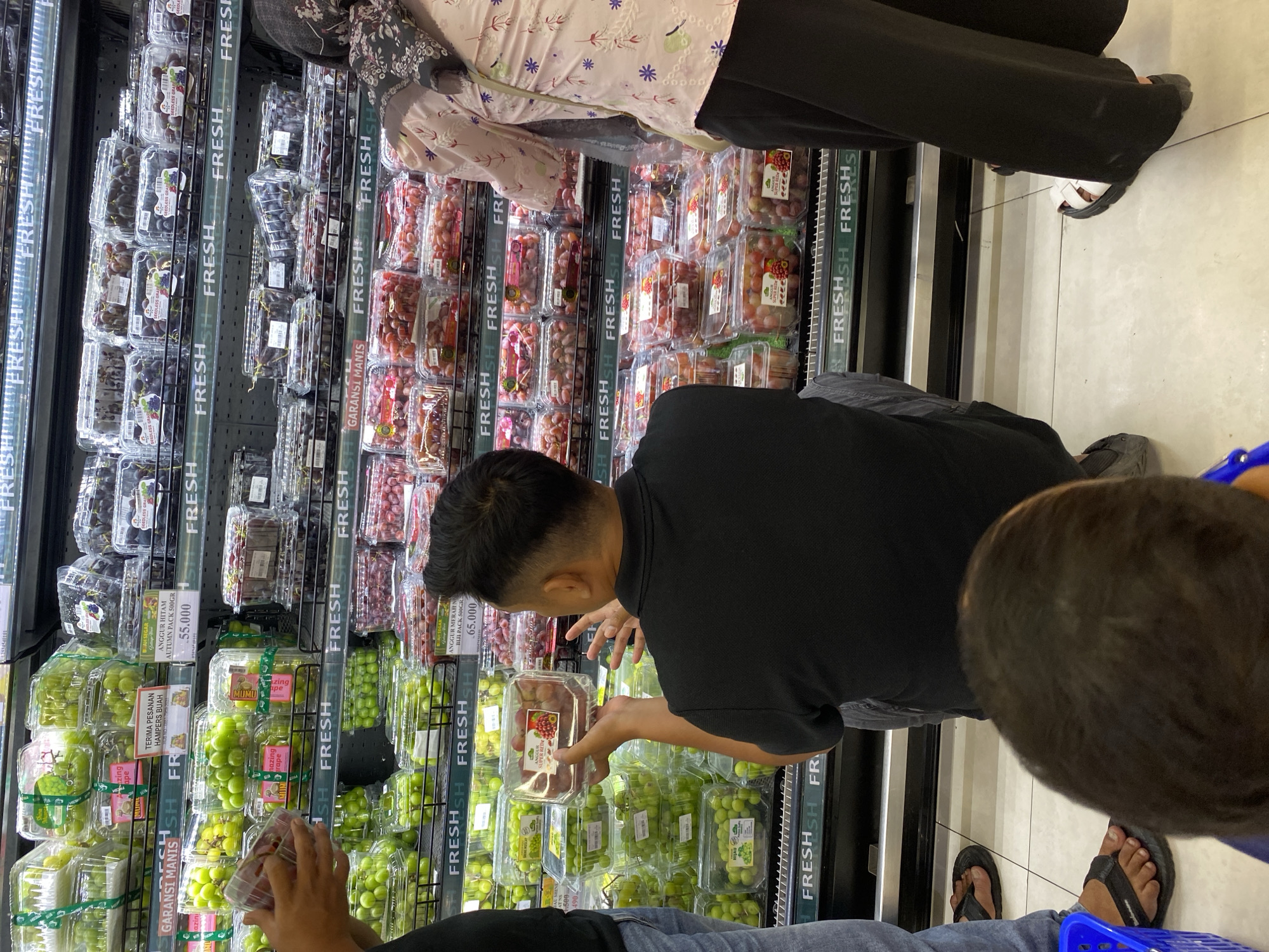 In a supermarket in Banda Aceh, Indonesia, a man is inspecting a clear plastic container of red grapes. He is wearing a black polo shirt and is viewed from behind. In front of him, a child's head is visible, also from behind. To the right, a woman in a pink floral shirt and black pants, wearing white sandals, stands with her back to the camera. To the left of the man, another person's legs, wearing grey jeans and black flip-flops, are visible. The background consists of refrigerated shelves filled with various types of grapes, predominantly green and red varieties, in clear plastic packaging. Signage with the word "FRESH" is visible on the shelves. The floor is tiled.