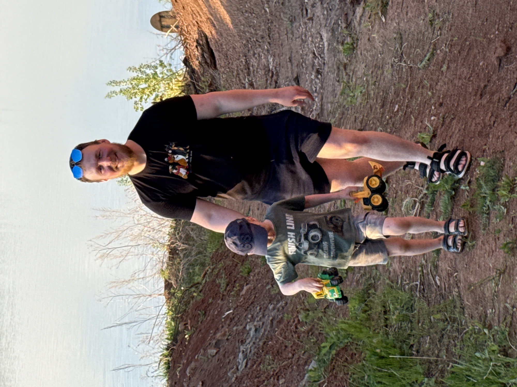 A fair-skinned adult male with a beard, wearing a black t-shirt, black shorts, and black and white sandals, stands next to a fair-skinned child wearing an olive green t-shirt, grey shorts, and black sandals. The male has blue sunglasses on his head. The child wears a dark cap. Both individuals are holding toy vehicles; the male holds a yellow and black toy tractor, and the child holds a green and yellow toy truck. They are standing on a dirt and grassy slope, with a body of water visible in the background. The location appears to be near a cliff or bank overlooking the water. The location may be in Прикамский, Russia.