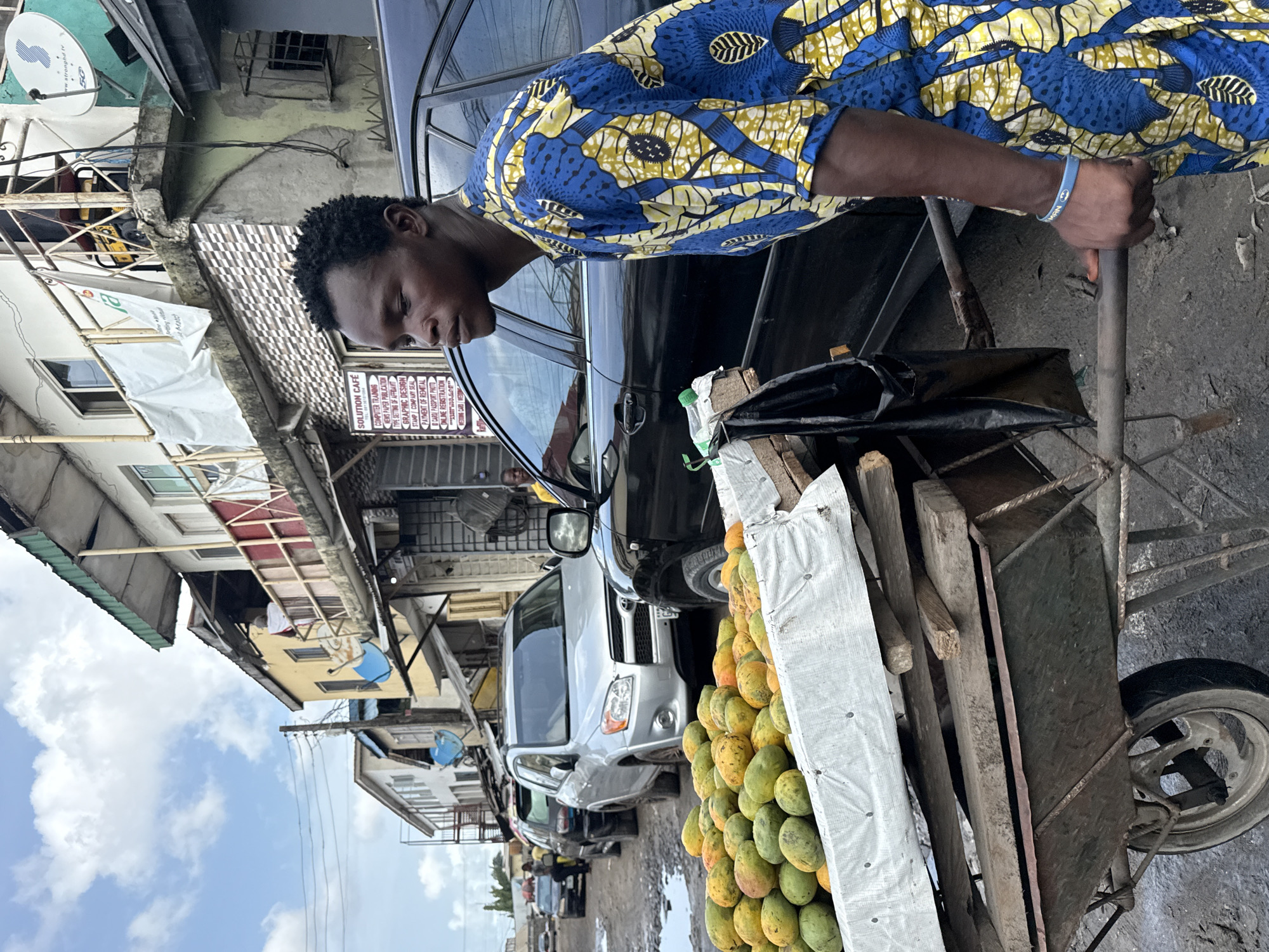 This media file presents a bustling daytime street scene in Lekki, Nigeria, centered around a young Black man vending fruit. The man, clad in a striking blue and yellow patterned shirt and a light blue bracelet, stands next to a black sedan, looking downwards, presumably attending to his goods or a customer.
His primary merchandise is displayed on a rustic, single-wheeled wooden pushcart brimming with ripe green and yellow mangoes. The background depicts a lively urban environment with various vehicles, including a silver SUV and other cars, along a street that appears to be unpaved in some sections. Buildings line the street, showcasing varied architectural styles and signs of age.
Notable details include satellite dishes on the buildings, one bearing the text "stronghd tv" and "5G." A prominent sign advertises "SOLUTION CAFÉ," offering services such as "COMPUTER TRAINING," "NEWSPAPER PUBLICATION," "GRAPHIC DESIGN," "STAMP COMPANY SEAL," "PAYMENT OF REMITTAL," "I CARD / PASSPORT PHOTO," and "ONLINE REGISTRATION." The clear sky with scattered white clouds indicates a sunny day. The scene captures an ordinary moment of commerce and daily life in a Nigerian urban setting.