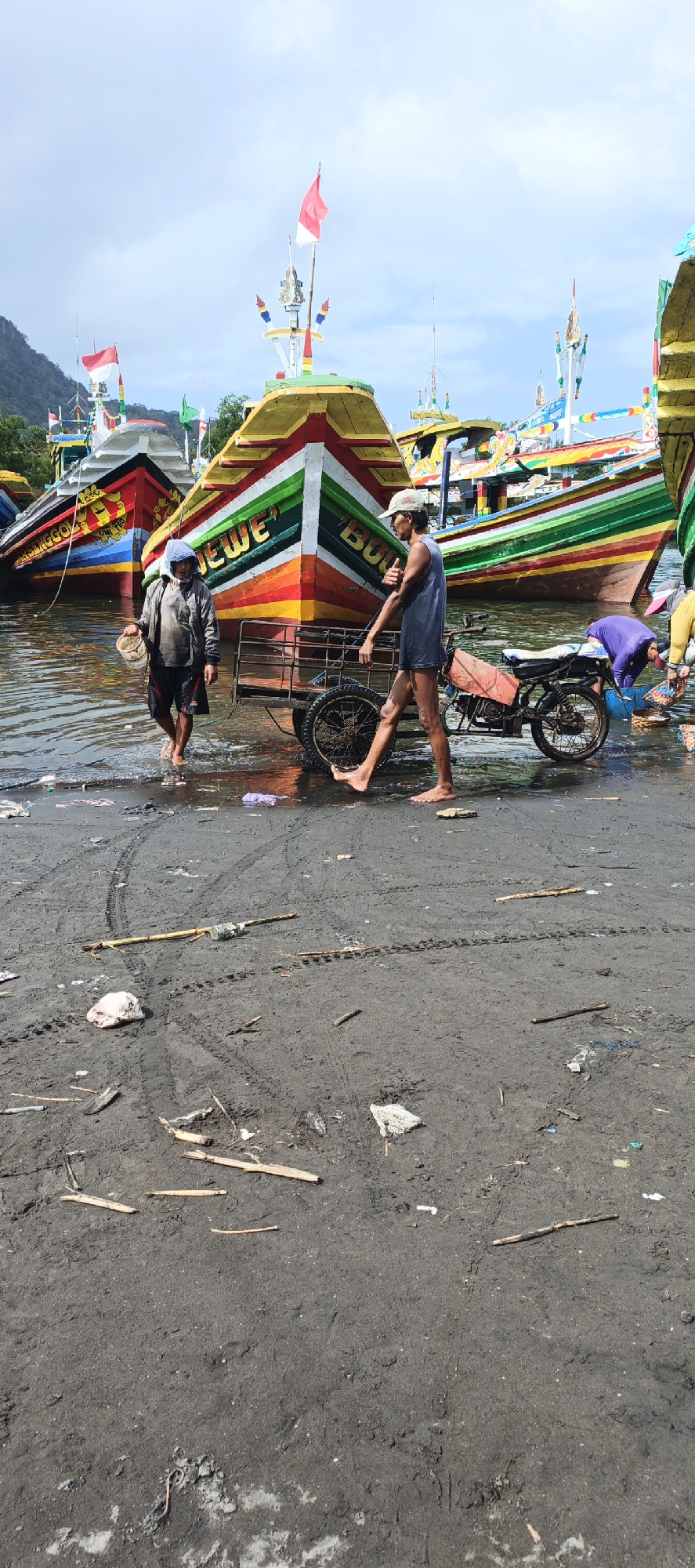 The image depicts a scene at the shore of the city of Puger, Indonesia, with several colorful fishing boats docked in the water. The foreground is a muddy, debris-strewn shoreline. In the middle ground, two men are present. One man, wearing a grey hooded jacket and dark shorts, walks through shallow water carrying a white bucket. Another man, wearing a dark sleeveless shirt, shorts, and a baseball cap, walks on the muddy shore, pulling a small cart attached to a motorcycle. The cart has a metal cage and one visible wheel. Behind the men, the brightly painted bows of several fishing boats are visible, adorned with intricate patterns and flags. The water appears calm. The sky is overcast with patches of blue.
