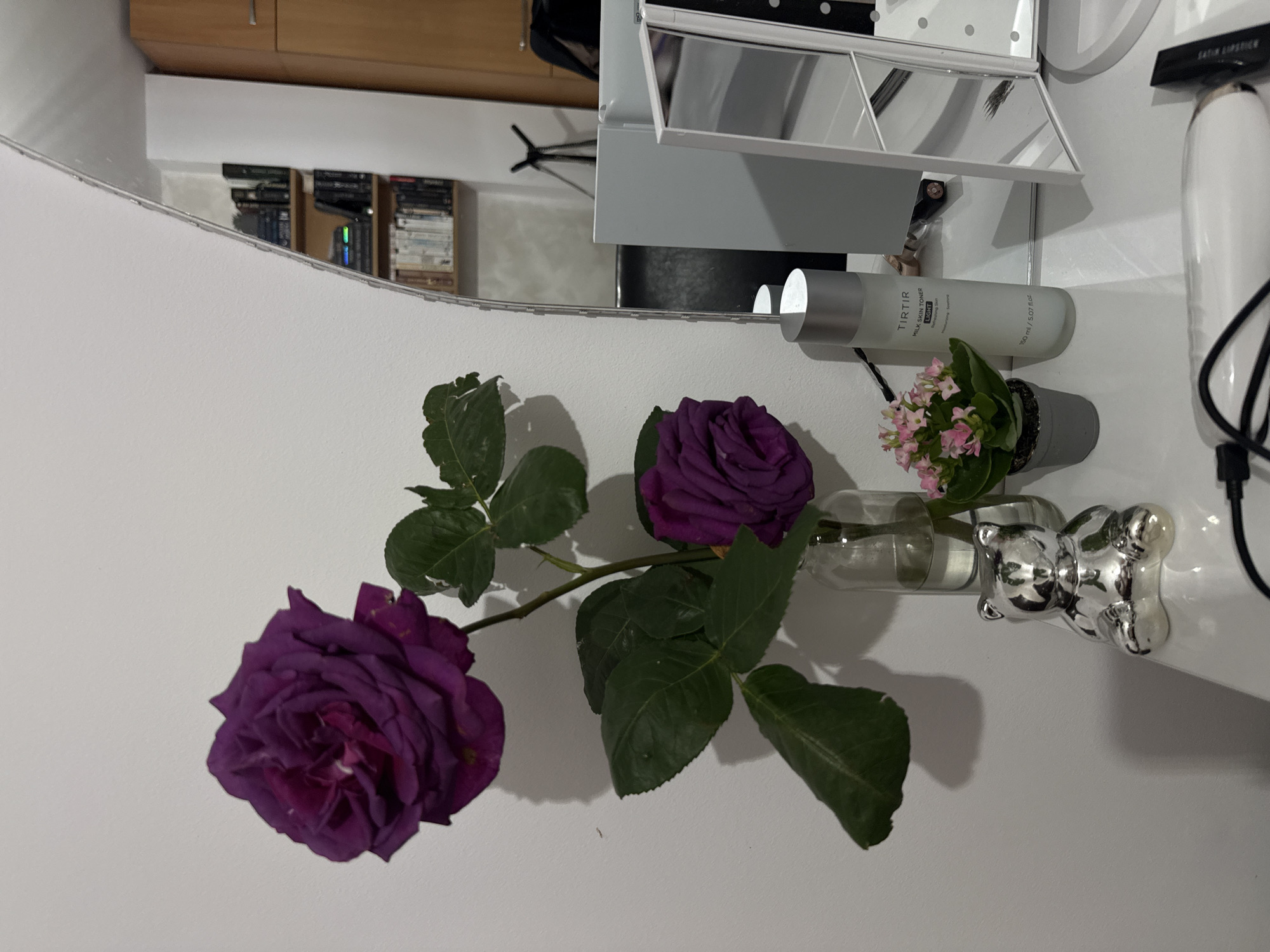 A close-up, slightly rotated, indoor shot shows a reflection in a mirror. In the foreground, a small glass vase holding two purple roses and a small potted pink flower arrangement rests on a white surface. Behind the vase are cosmetic items, including a white bottle labeled "SATIN LIPSTICK" and a gray bottle labeled "TIRTIR MILK SKIN TONER". The mirror reflects a room with a shelf of books and a lighting fixture. The location is not explicitly stated, and there is no information in the image that could positively identify the location as Blagoevgrad, Bulgaria.