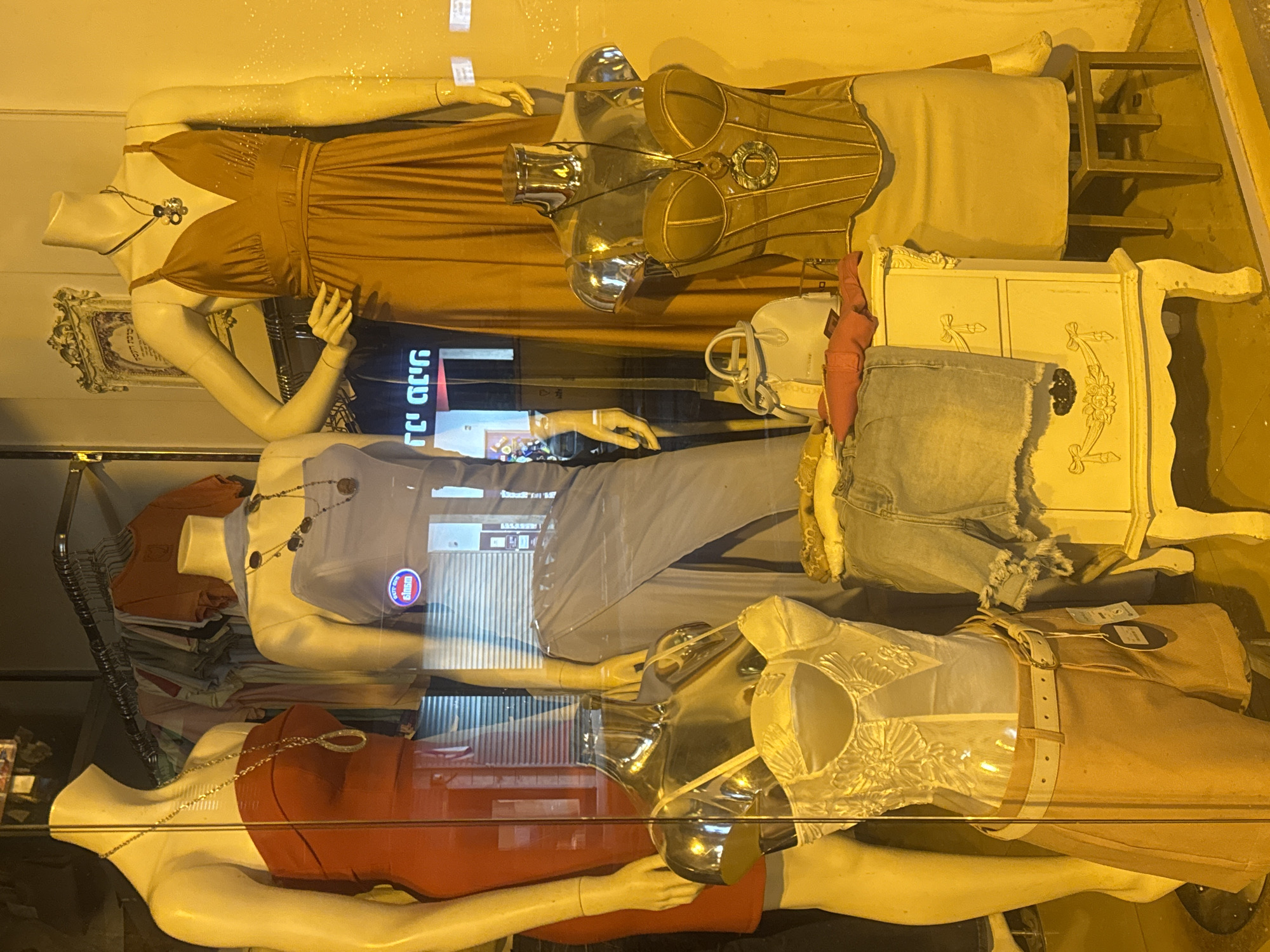 A clothing store display window in Ramat Gan, Israel, features three mannequins dressed in different outfits.
The top mannequin wears a long, brown dress with a necklace. A gold-colored corset is displayed over the dress. An antique-style white dresser stands next to the mannequin.
The middle mannequin wears a light purple dress with a necklace. A pile of folded clothes are on the left.
The bottom mannequin wears a red dress with a chain necklace.
The display window has reflections visible in it, suggesting a street scene outside.