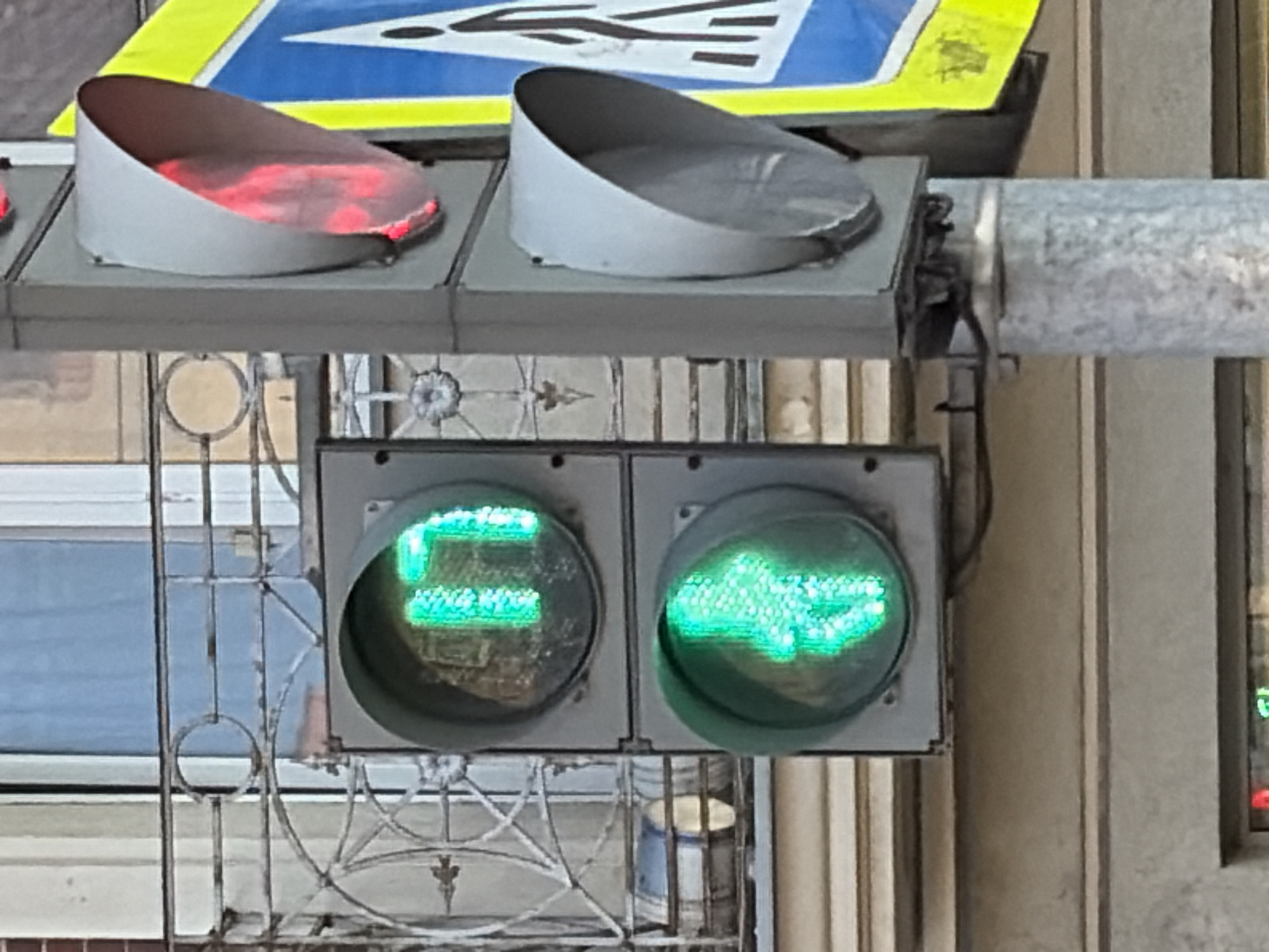 In the city of Unknown, Unknown, a close-up, eye-level shot focuses on a traffic signal pole. The pole is metallic and appears to be a light gray color. Attached to the pole are two traffic light units, one above the other, both emitting green light. The upper traffic light displays a green "m" or "w" shape formed by LED lights, while the lower traffic light shows a green silhouette of a walking person. To the right of the traffic lights, a bright yellow and blue pedestrian crossing sign is visible. The background includes a building with a decorative metal railing on a balcony and a window reflecting the sky. The image is taken in daylight.