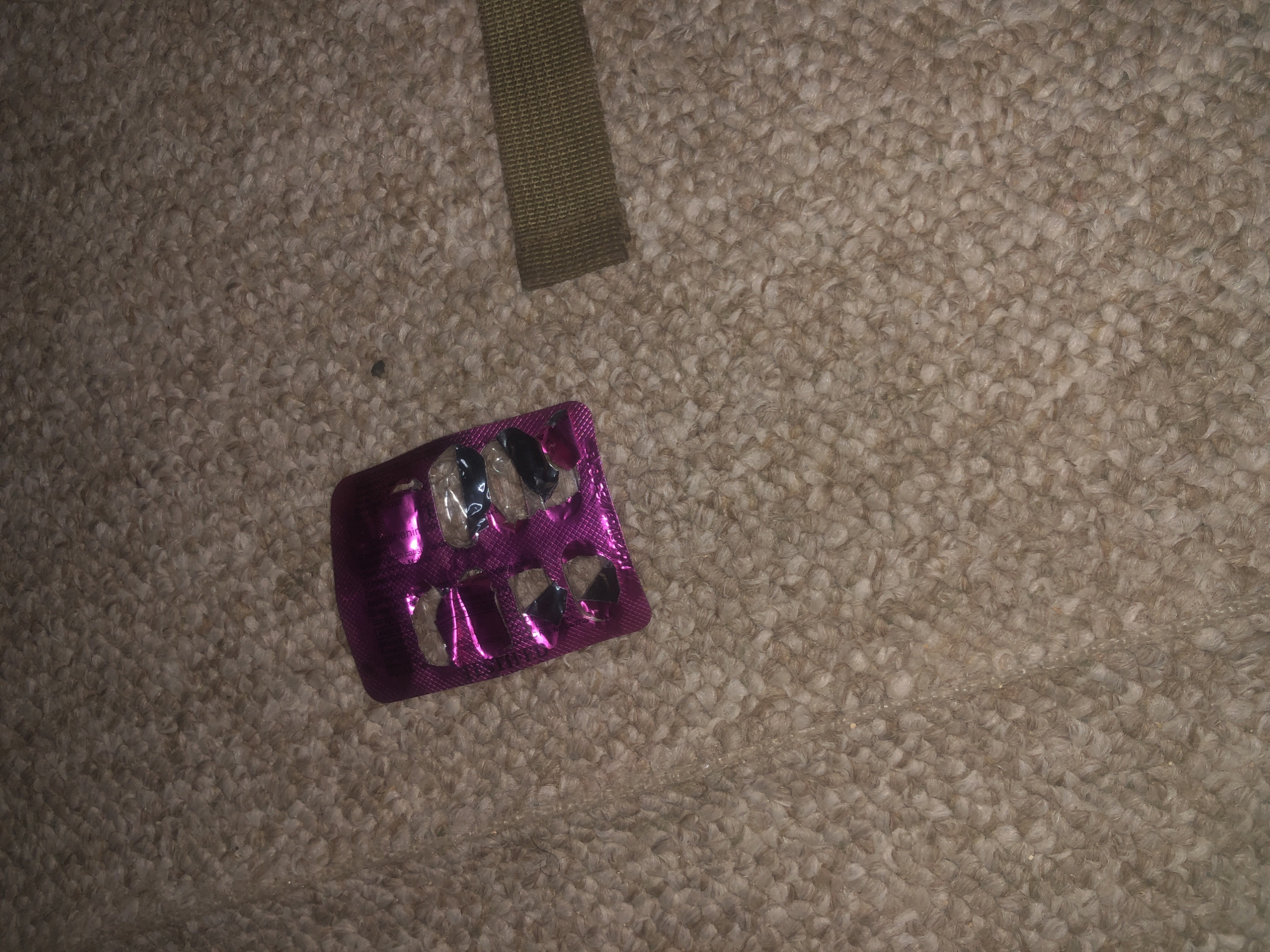 A pink blister pack of pills, with some pills missing, lies on a beige carpet. To the right of the blister pack is a strip of olive green fabric, possibly part of a belt. The lighting is dim and the image is slightly blurry. The scene is indoors, on the floor of a room, but there is no indication of it being in Jalingo, Nigeria or any specific city.