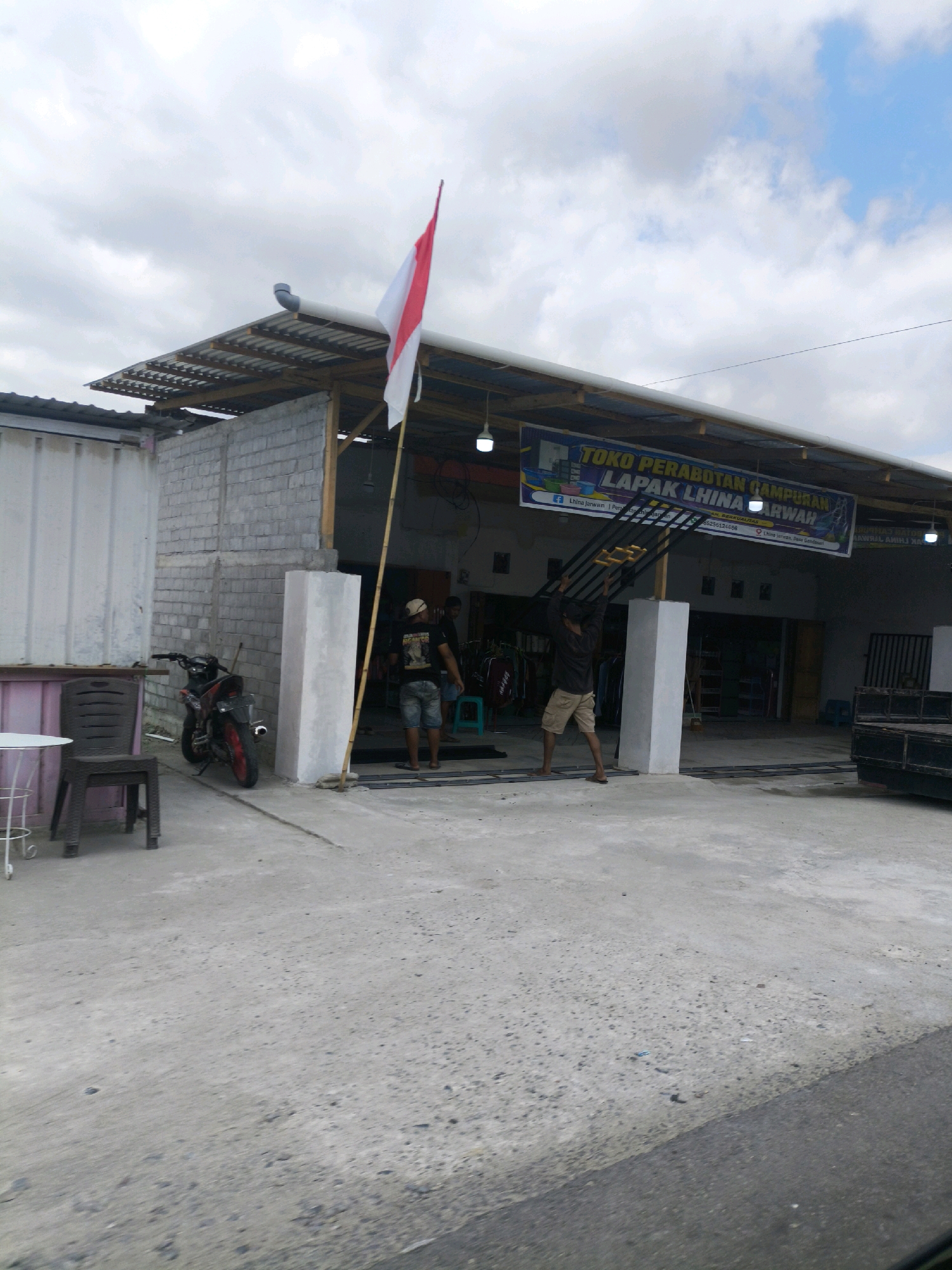 The image shows a street view in Diloniyohu, Indonesia. A storefront with a corrugated metal roof stands on the right side of the image. A banner above the entrance reads "TOKO PERABOTAN CAMPURAN LAPAK LHINA AMIRWAH". Two men are standing in front of the store. One man on the left is wearing a black t-shirt and denim shorts, and the other man on the right is wearing a black t-shirt and khaki shorts. A motorcycle is parked to the left of the store entrance. In the foreground, a concrete surface stretches across the image, with a section of asphalt road visible on the right. The sky is cloudy, with patches of blue sky visible.